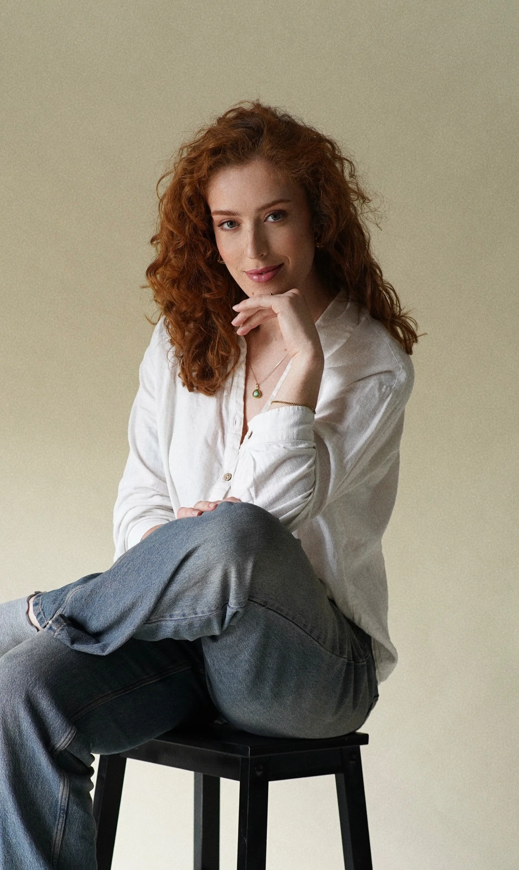 A woman with red curly hair sitting on a black stool, wearing a white shirt and blue jeans, looking at the camera with her chin resting on her hand.