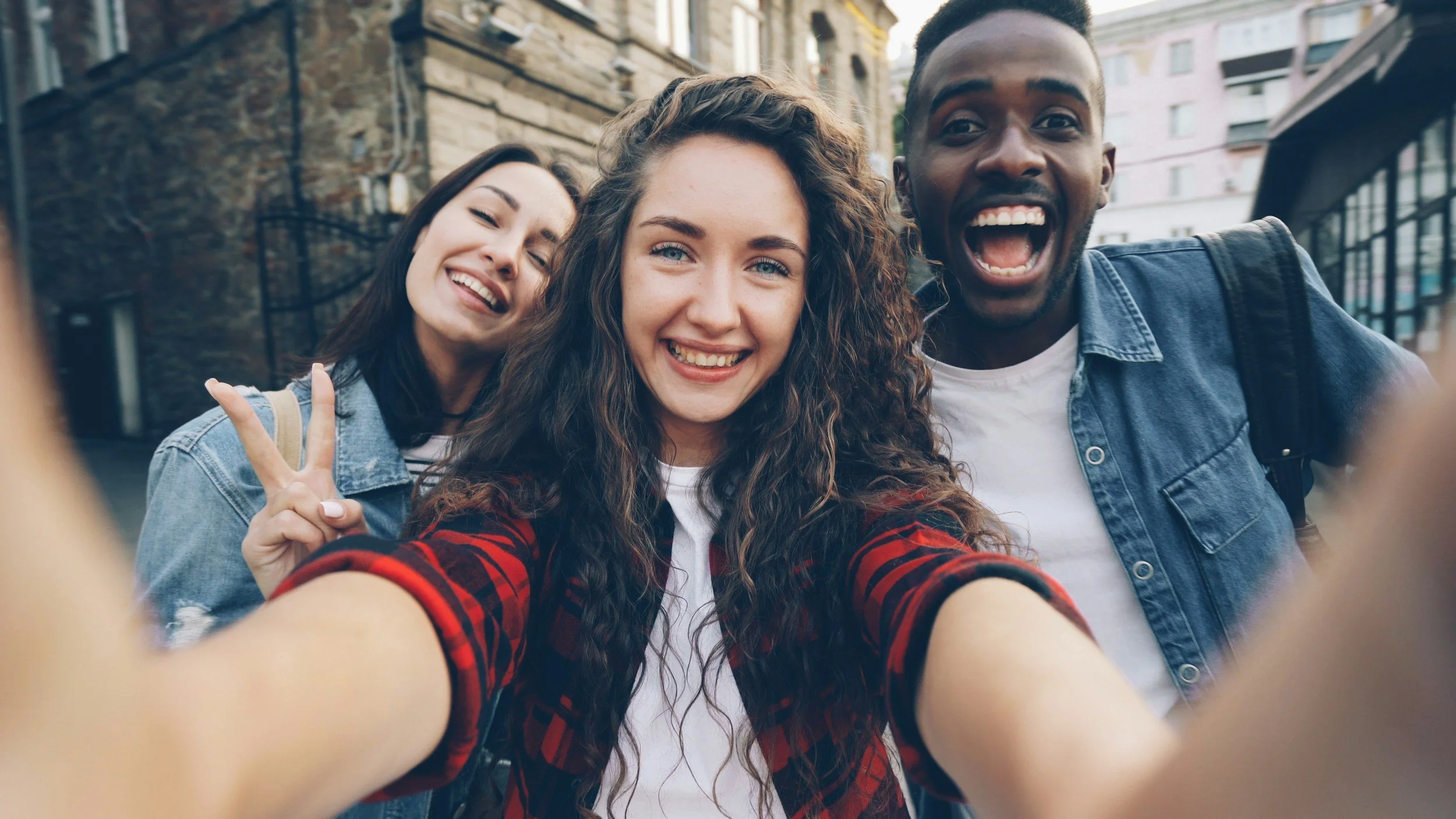 Three young friends taking a selfie outdoors. The girl in the center is smiling, with curly hair and wearing a red and black checkered shirt. The girl on the left is smiling and making a peace sign, and the guy on the right is smiling widely with his mouth open and wearing a denim jacket.