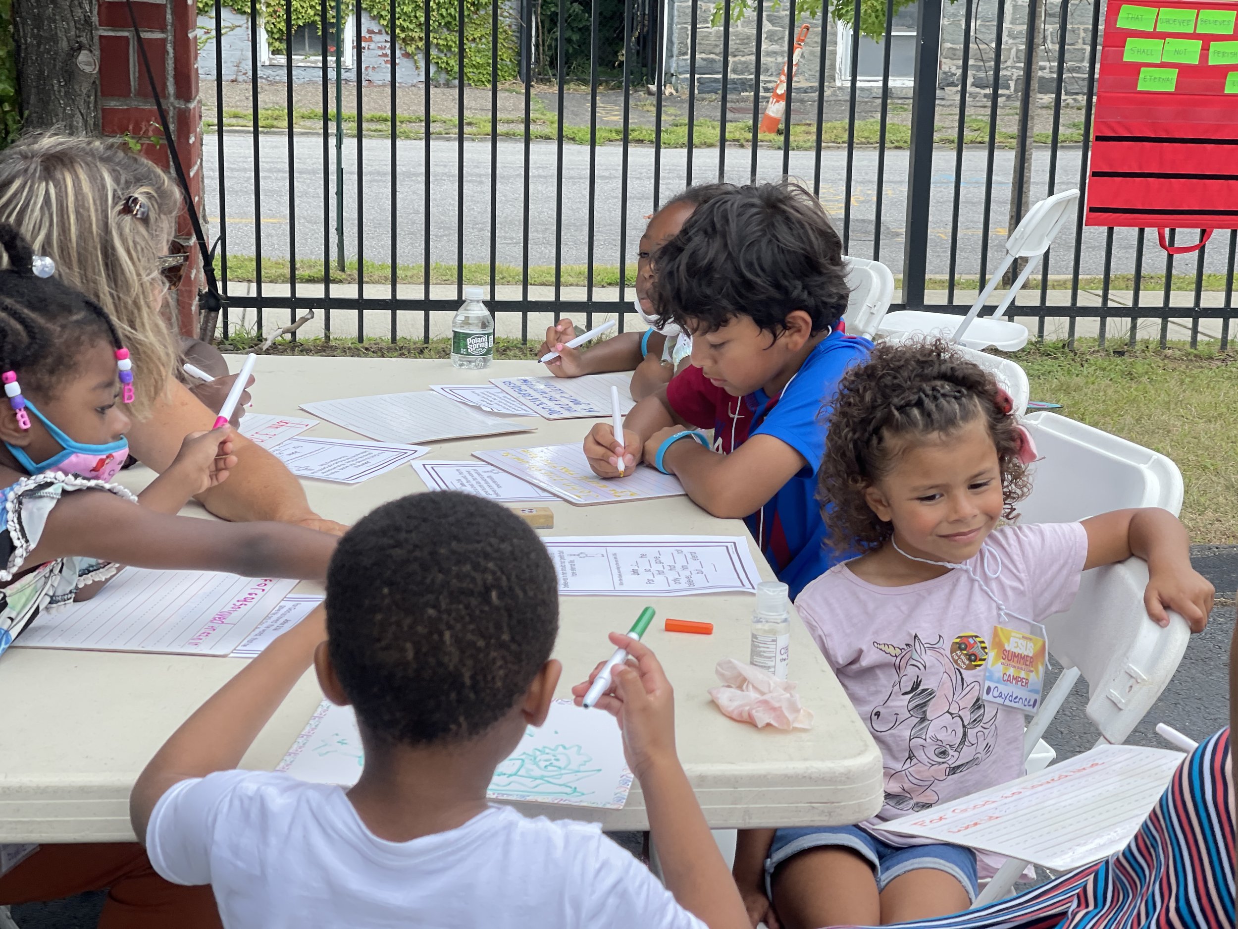 Children and an adult sitting outdoors at a table, engaged in drawing or writing activities, with papers, markers, and a water bottle on the table, behind a fence.