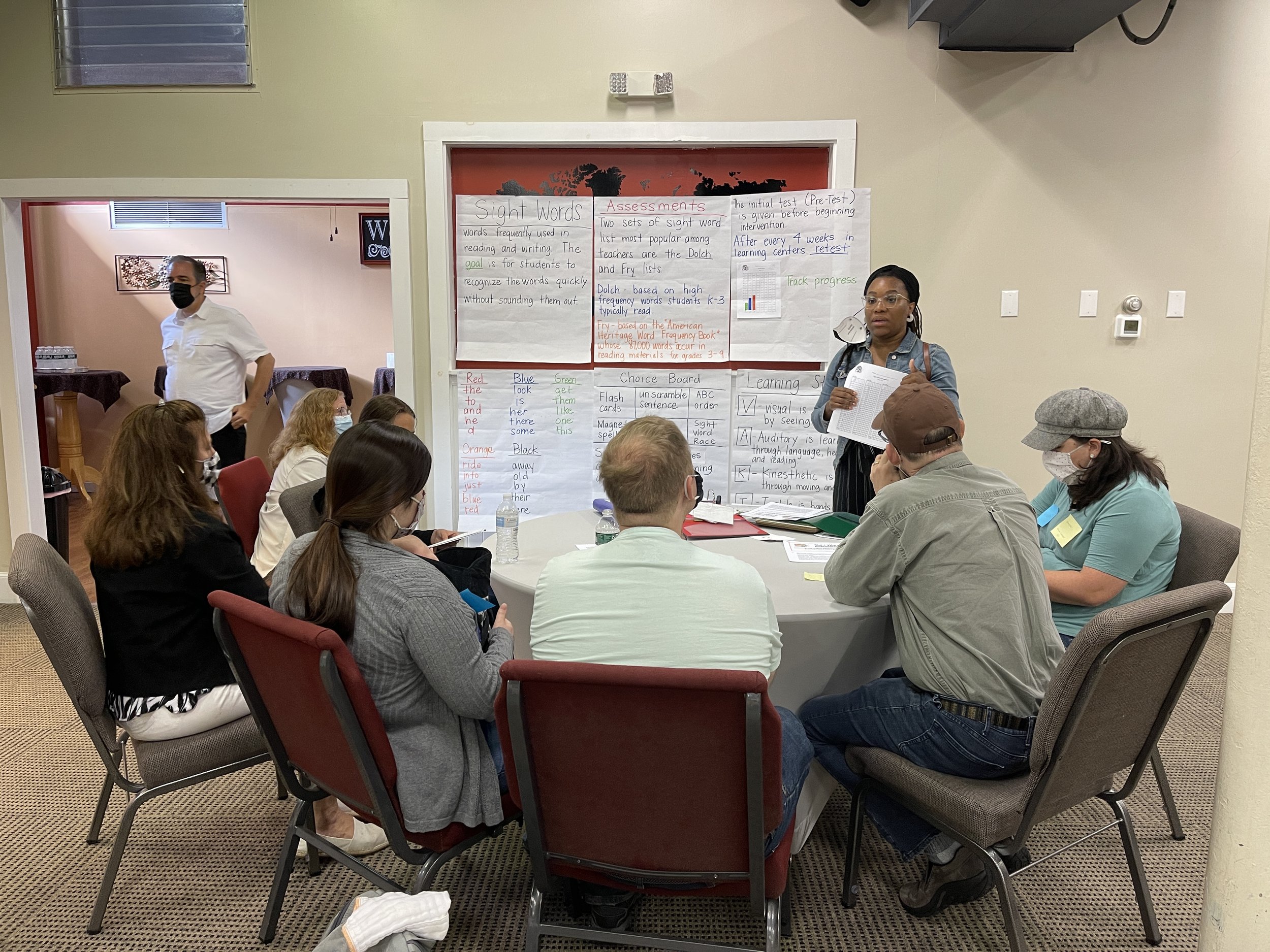 A group of adults sitting around a round table in a classroom or workshop setting. A woman is standing near a whiteboard presenting, while others are seated with notebooks, water bottles, and masks. The whiteboard contains written notes and charts related to assessment and reading skills.