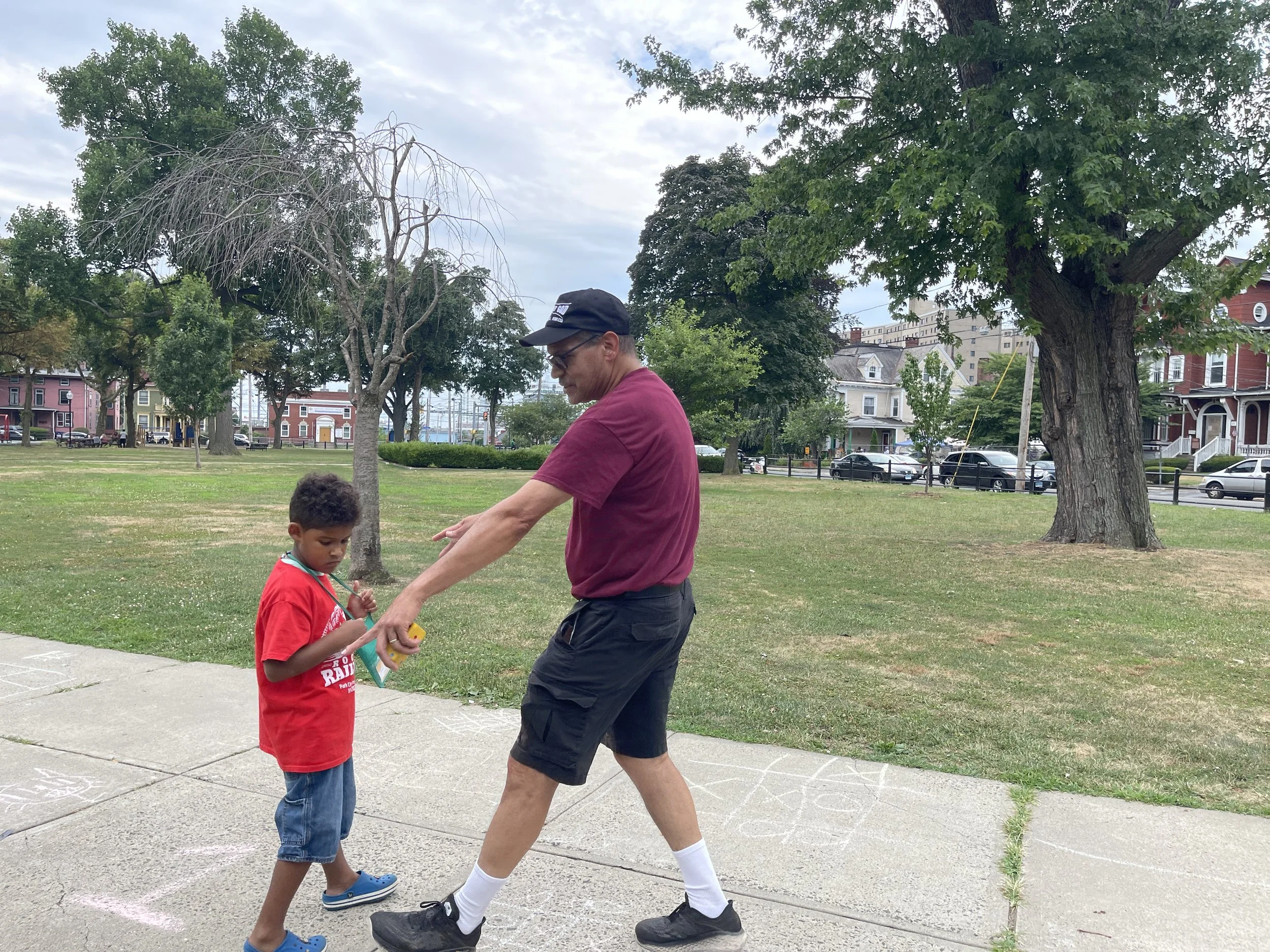 A man and a young boy are playing hopscotch on a sidewalk in a park. The man is wearing a maroon t-shirt, black shorts, and a black cap. The boy is in a red t-shirt, jeans, and blue sandals. Trees and houses are visible in the background.