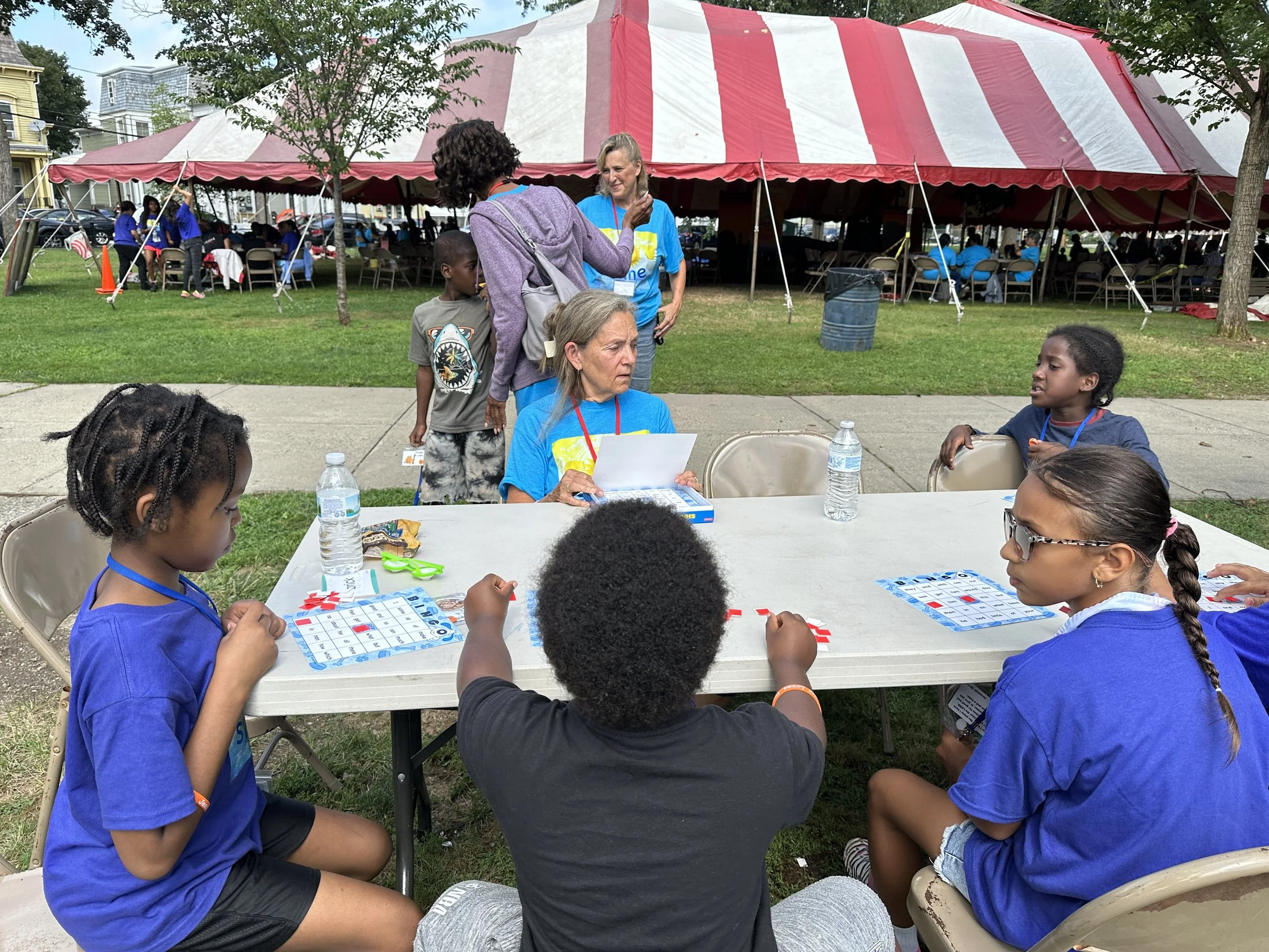 Children playing Bingo at an outdoor event with tents, tables, and people in the background.