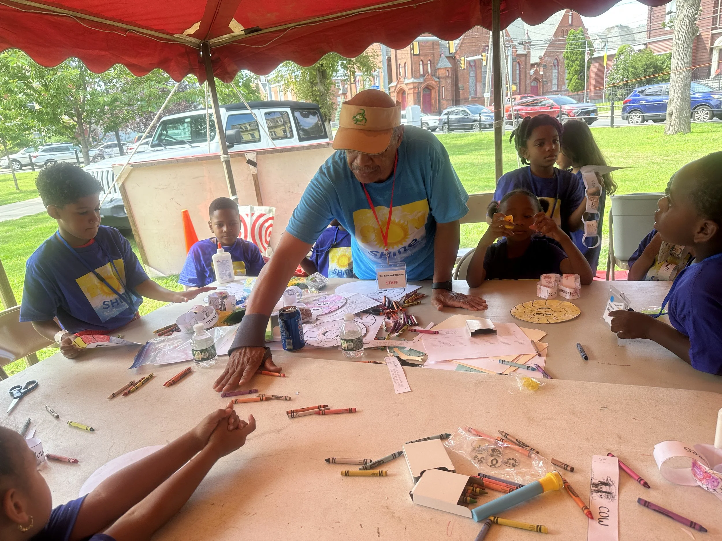 A group of children and an adult participating in an outdoor arts and crafts activity under a red canopy. The adult, wearing a cap and a blue t-shirt, is leaning over a table filled with crayons, paper, and craft supplies. The children are engaged, s