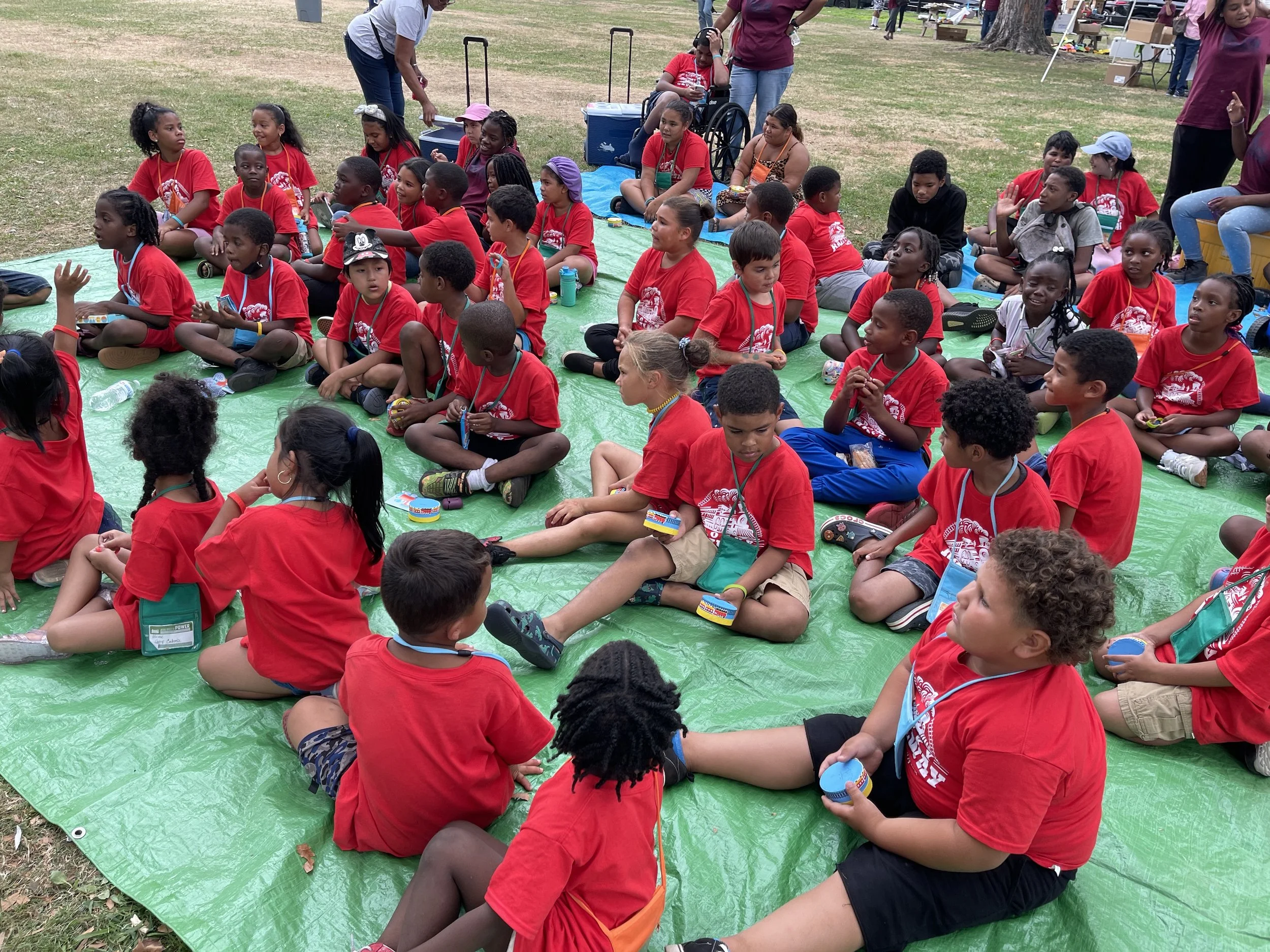 Group of children sitting on green tarp outdoors during a group event or picnic, all wearing red t-shirts, with some adults standing around.
