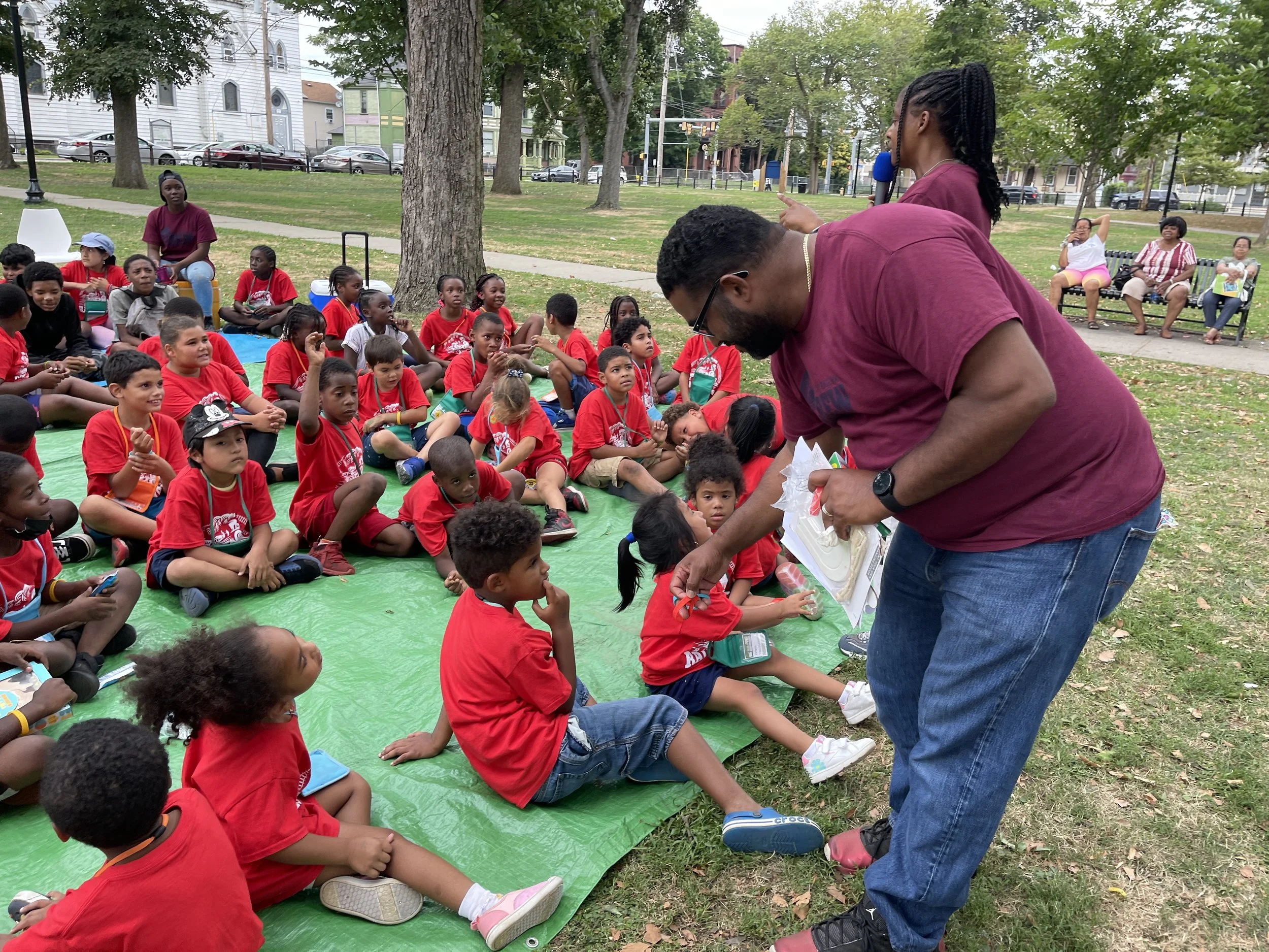 A group of children in red shirts sitting on a green tarp in a park, listening attentively to a man in a maroon shirt and blue jeans, who is handing something to a young girl. Several adults are sitting on benches in the background, and there are tre