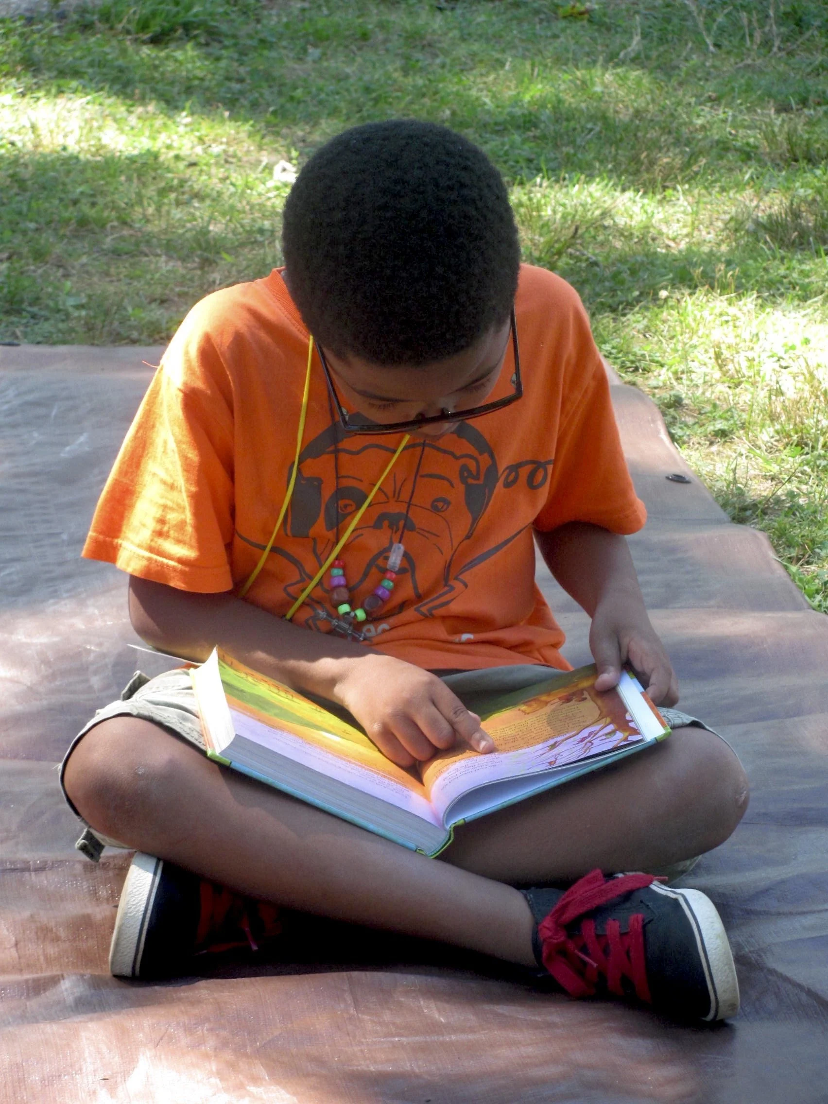 A boy sitting cross-legged outdoors on a brown tarp, reading a colorful book.