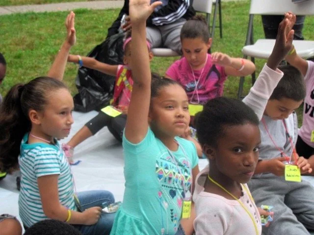 Group of children sitting on the ground during an outdoor activity, with one girl raising her hand.