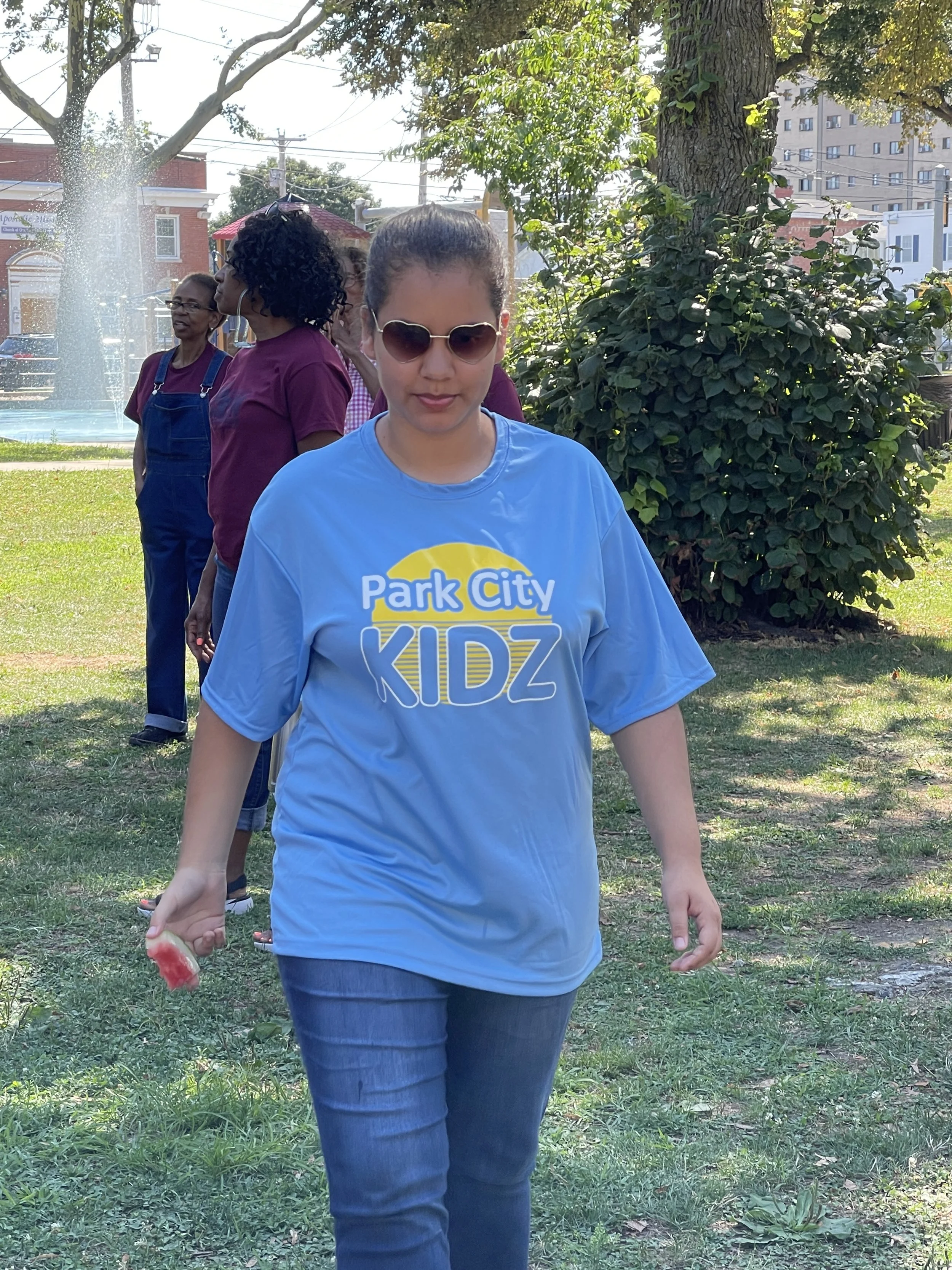 Woman walking outdoors in park, wearing sunglasses and a blue 'Park City KIDZ' t-shirt, with girls in maroon shirts and overalls standing in the background near trees.