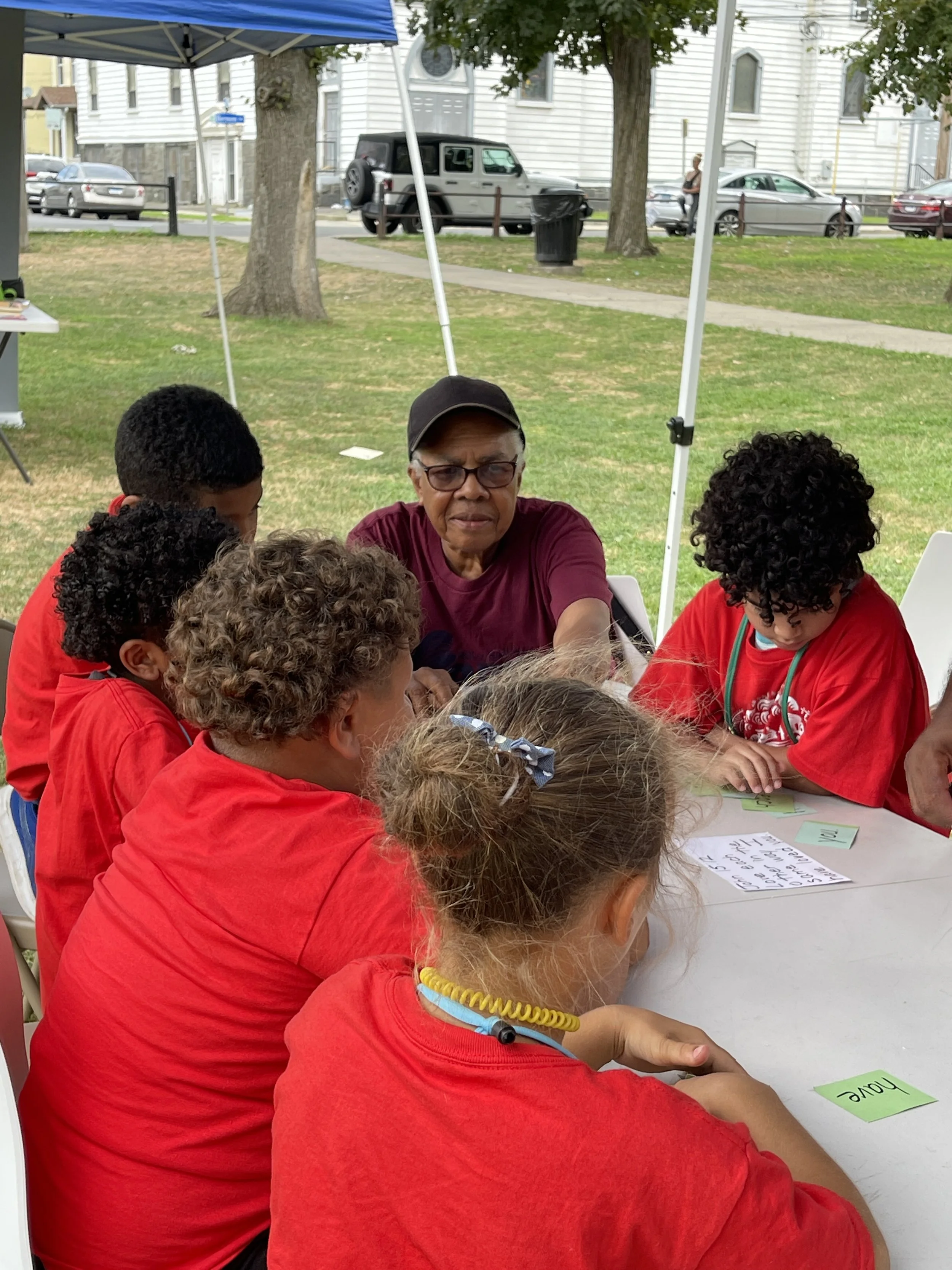 An elderly woman and six children gathered around a table outdoors, working on educational activities. The children are wearing red shirts, and the woman is wearing glasses, a dark cap, and a maroon shirt. There is a white piece of paper with writing