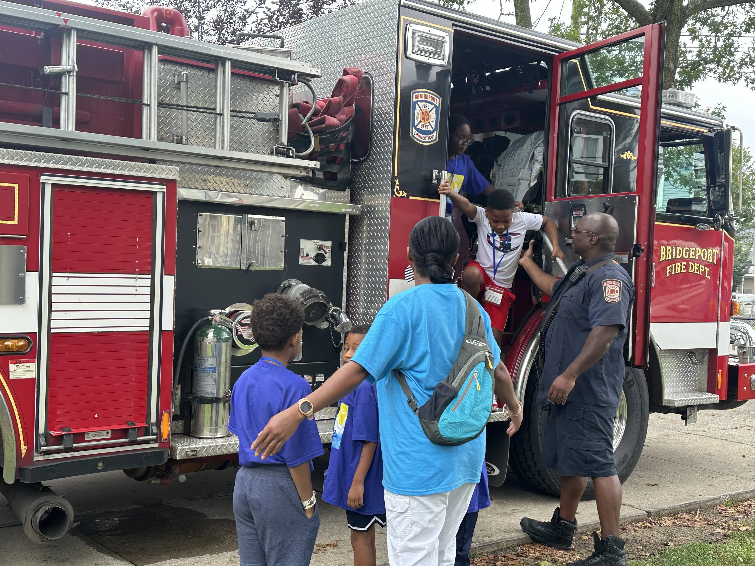 A group of children and adults gathered around a red fire truck from the Bridgeport Fire Department. One child is climbing into the truck with an adult assisting, while others are observing nearby.