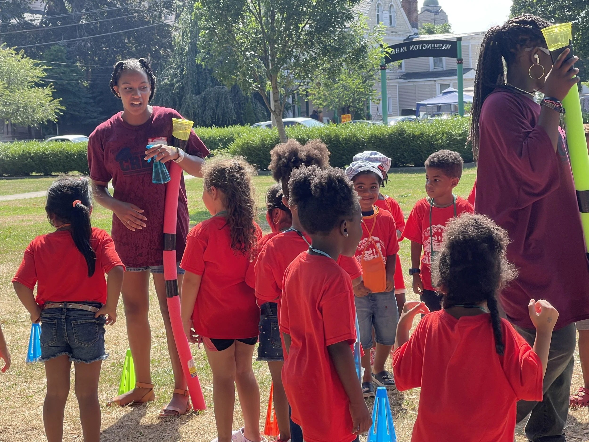 Children participating in an outdoor group activity on a sunny day, with instructors guiding them, set in a grassy area with trees, bushes, and buildings in the background.