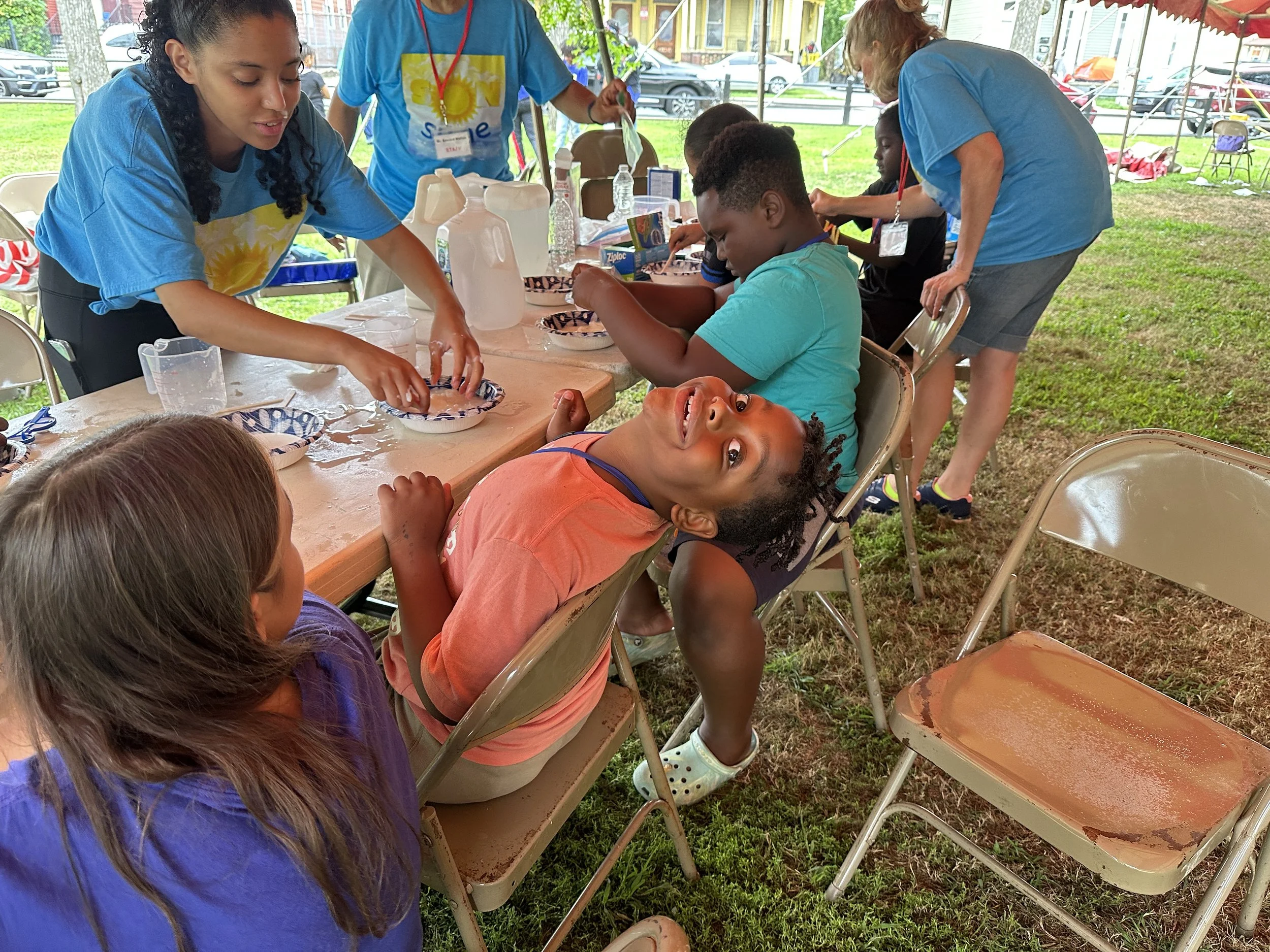 Children and adults participating in a craft activity outdoors under a tent, with some seated at a long table and others standing around, on a grassy area with chairs and tents in the background.