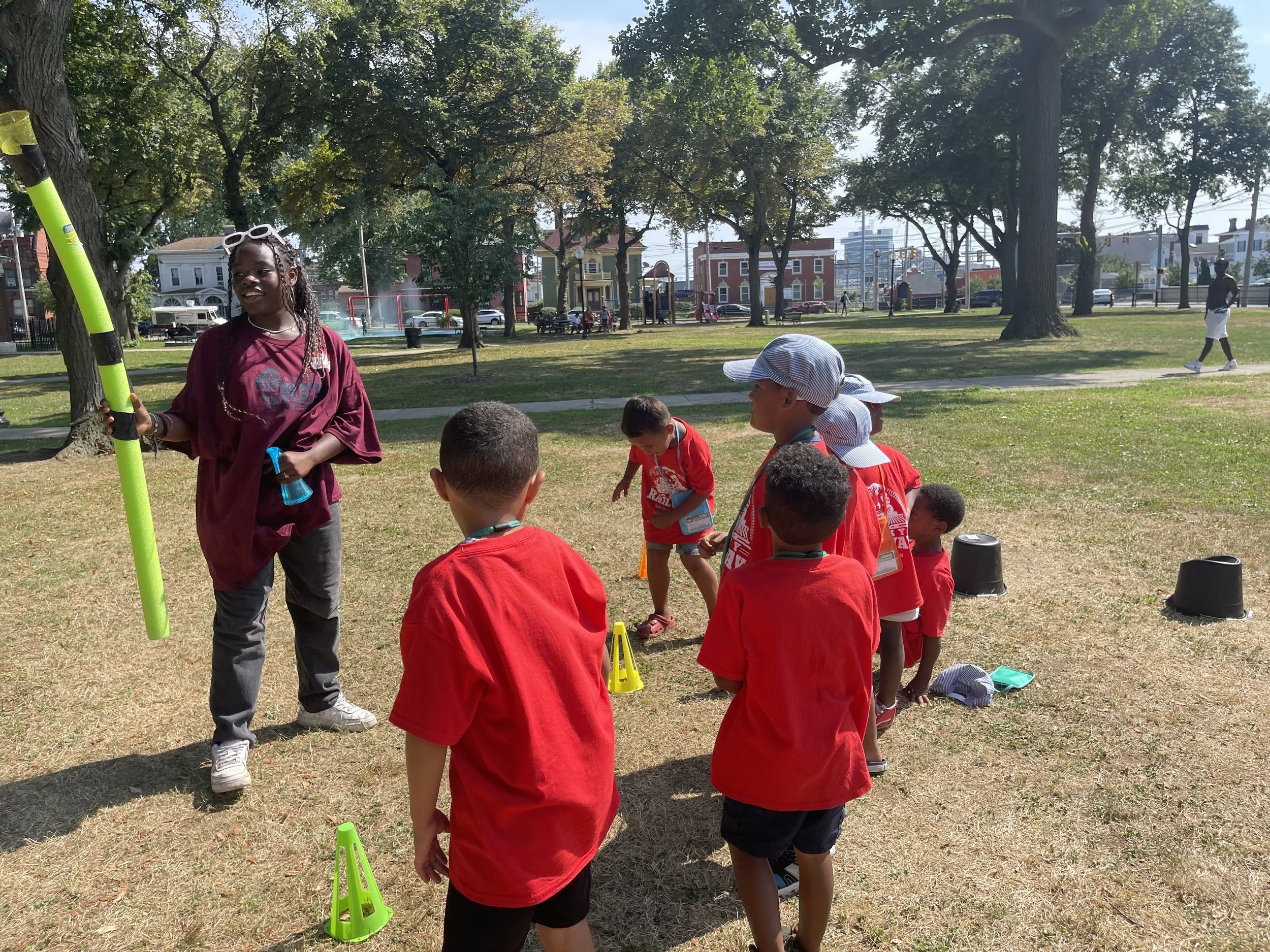 Children wearing red shirts standing in a line outdoors on grass with a woman holding a long yellow and black object, possibly a bat, in a park with trees, houses, and a playground in the background.