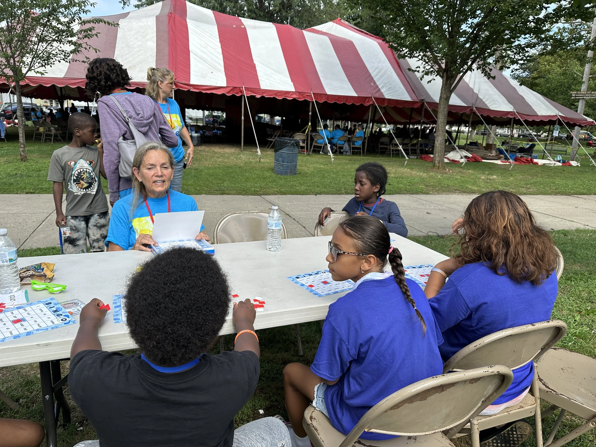 People gathered outdoors, some sitting at a folding table playing bingo with game cards and markers, others standing and talking near a large red and white striped event tent, possibly at a community or festival event.
