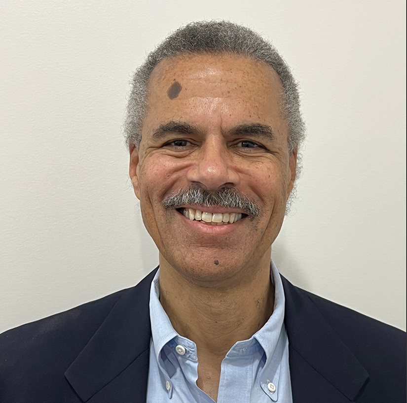 A smiling middle-aged man with gray curly hair, a mustache, and a small dark mole on his forehead. He's wearing a black blazer over a light blue button-up shirt and standing against a plain white wall.
