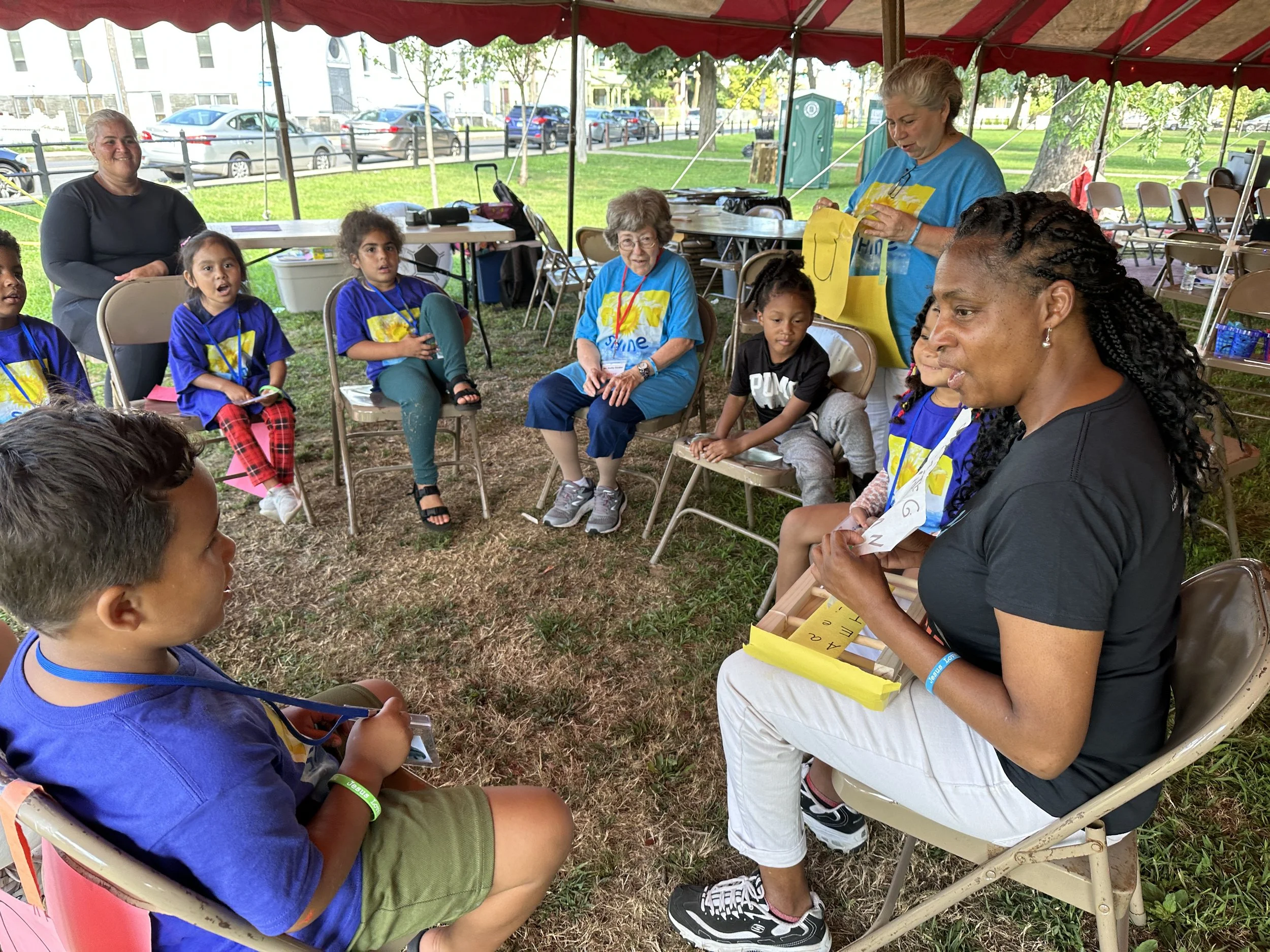 Children sitting in a semicircle under a tent while a woman with curly hair teaches them at an outdoor event.