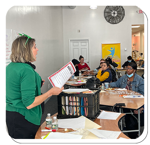 A woman standing in front of a classroom holding a paper, appearing to teach or present to a group of students seated at tables.