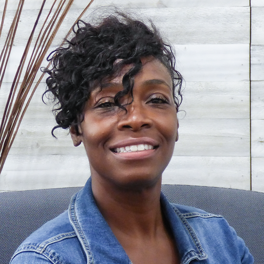 A smiling woman with short curly black hair, wearing a denim jacket, sitting in front of a wooden wall.
