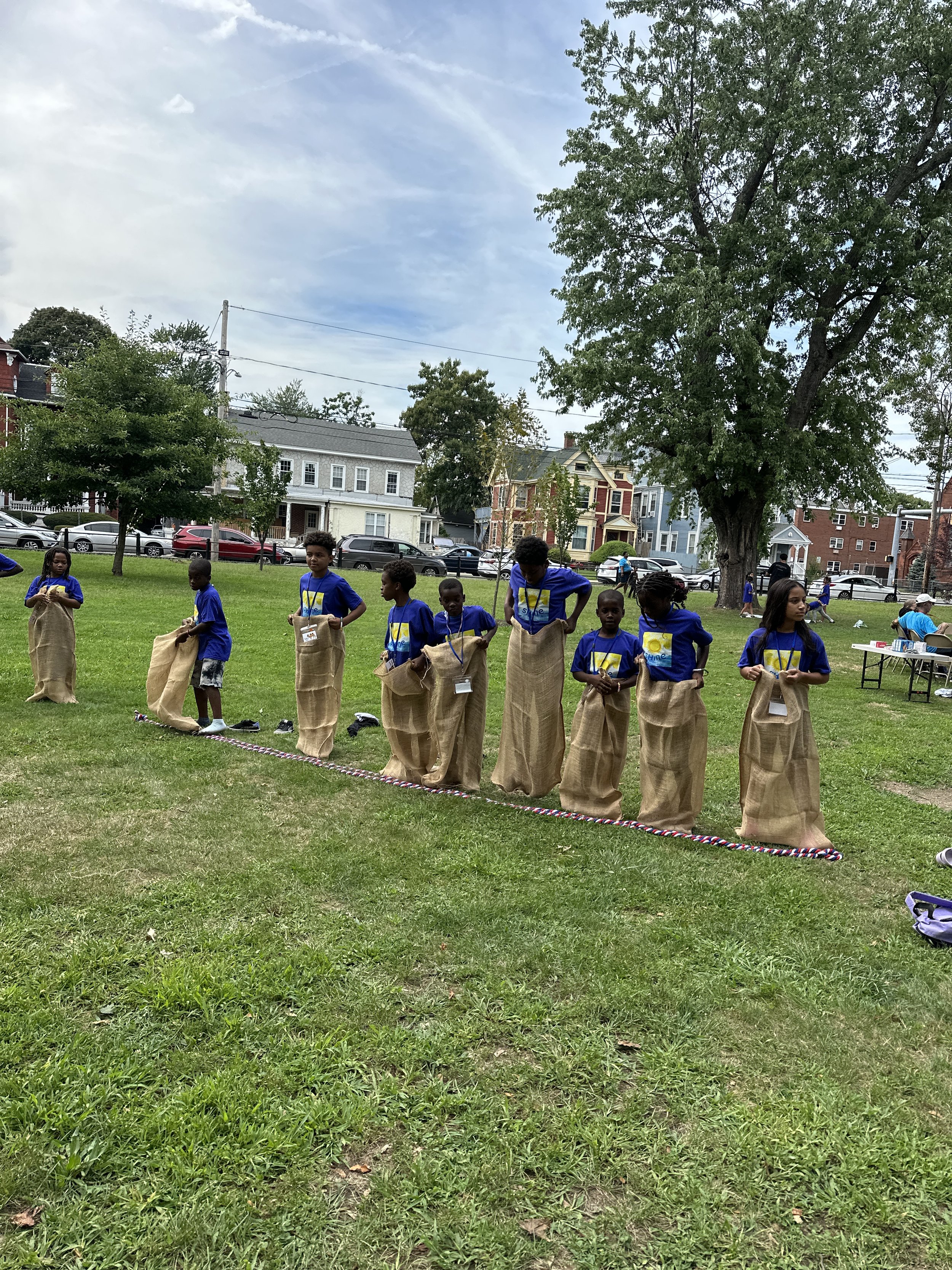 Children participating in a sack race outdoors on a grassy field. They are lined up with sacks, ready to race, wearing blue shirts.