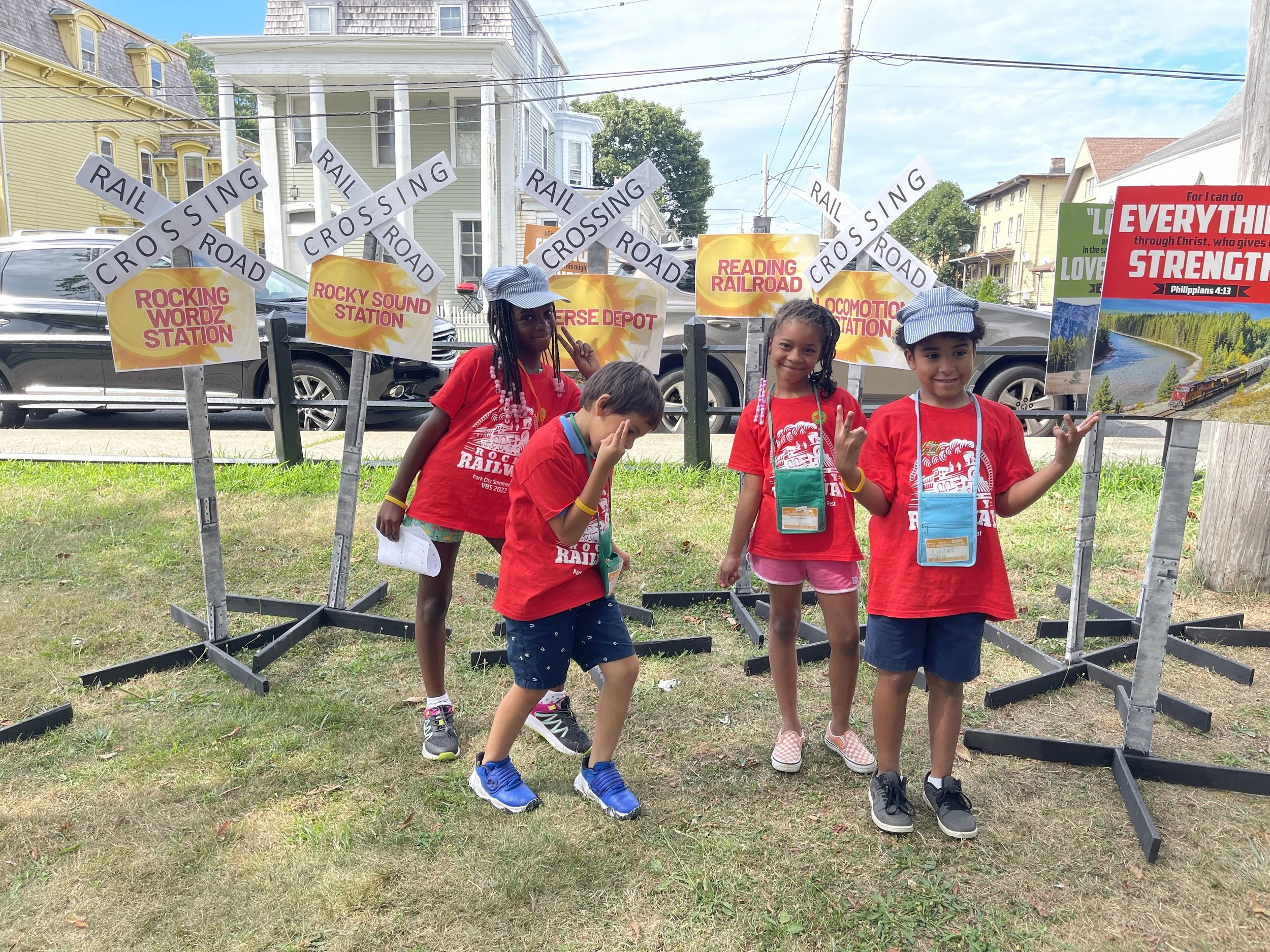 Four children wearing red manning a train crossing themed booth with signs reading 'Crossing Road,' 'Rail Crossing,' and stations named 'Rocking Wordz Station,' 'Rocky Sound Station,' 'Diverse Depot,' and 'Locomotion Station' in a park area on a sunn