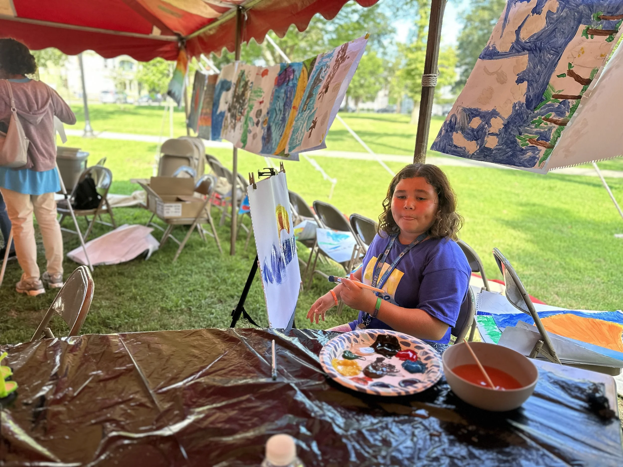 A young girl with curly hair and a purple T-shirt sitting at a table under a red canopy, painting colorful pictures. Several completed paintings are hanging above her. The table has a paint palette, a bowl of red paint, and a black plastic sheet cove