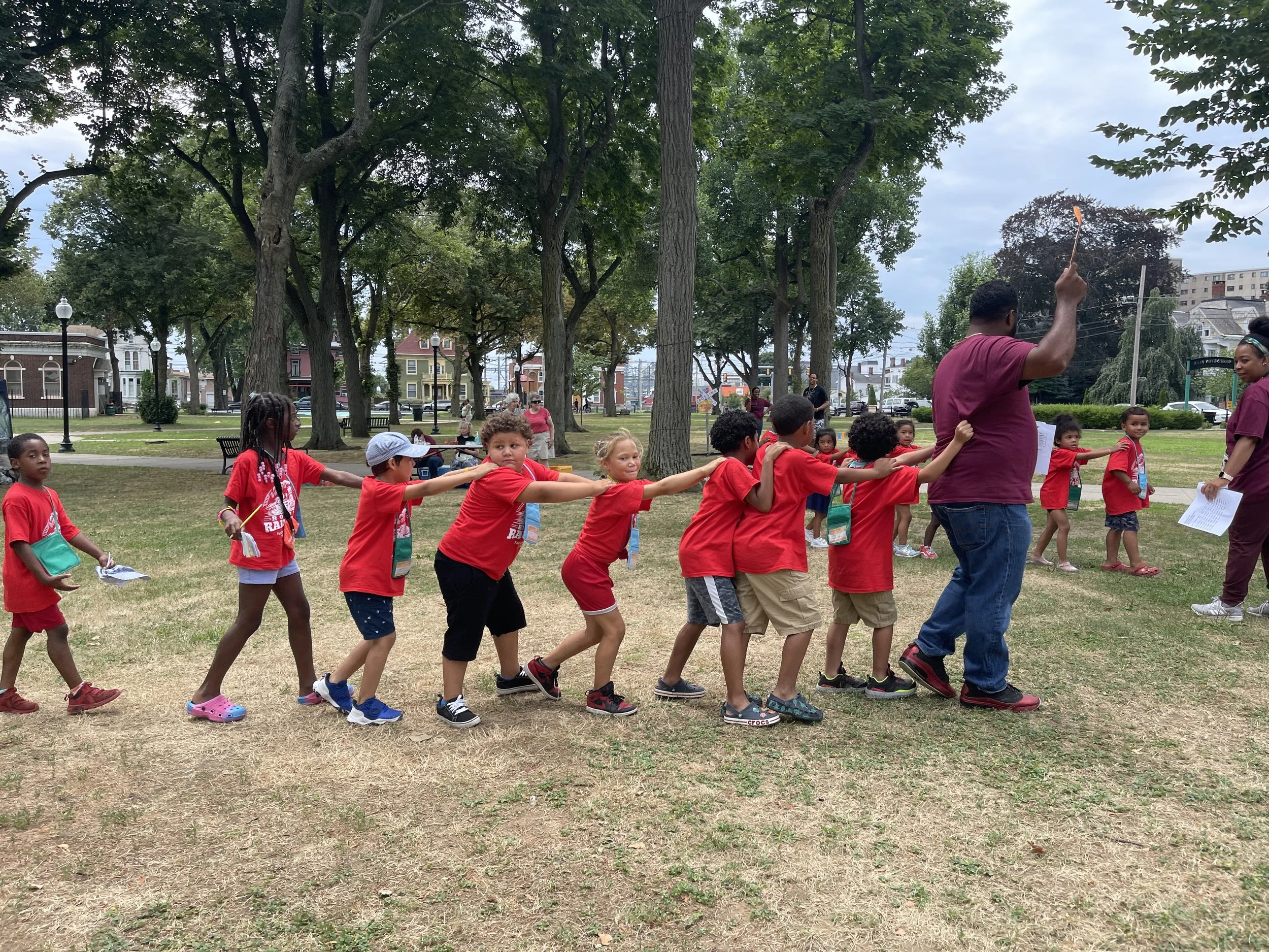 Children in red shirts forming a conga line in a park, participating in an outdoor group activity.