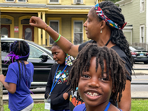 A woman with braided hair and a colorful headband is smiling and raising her arm, surrounded by three children outdoors in a neighborhood on a cloudy day.