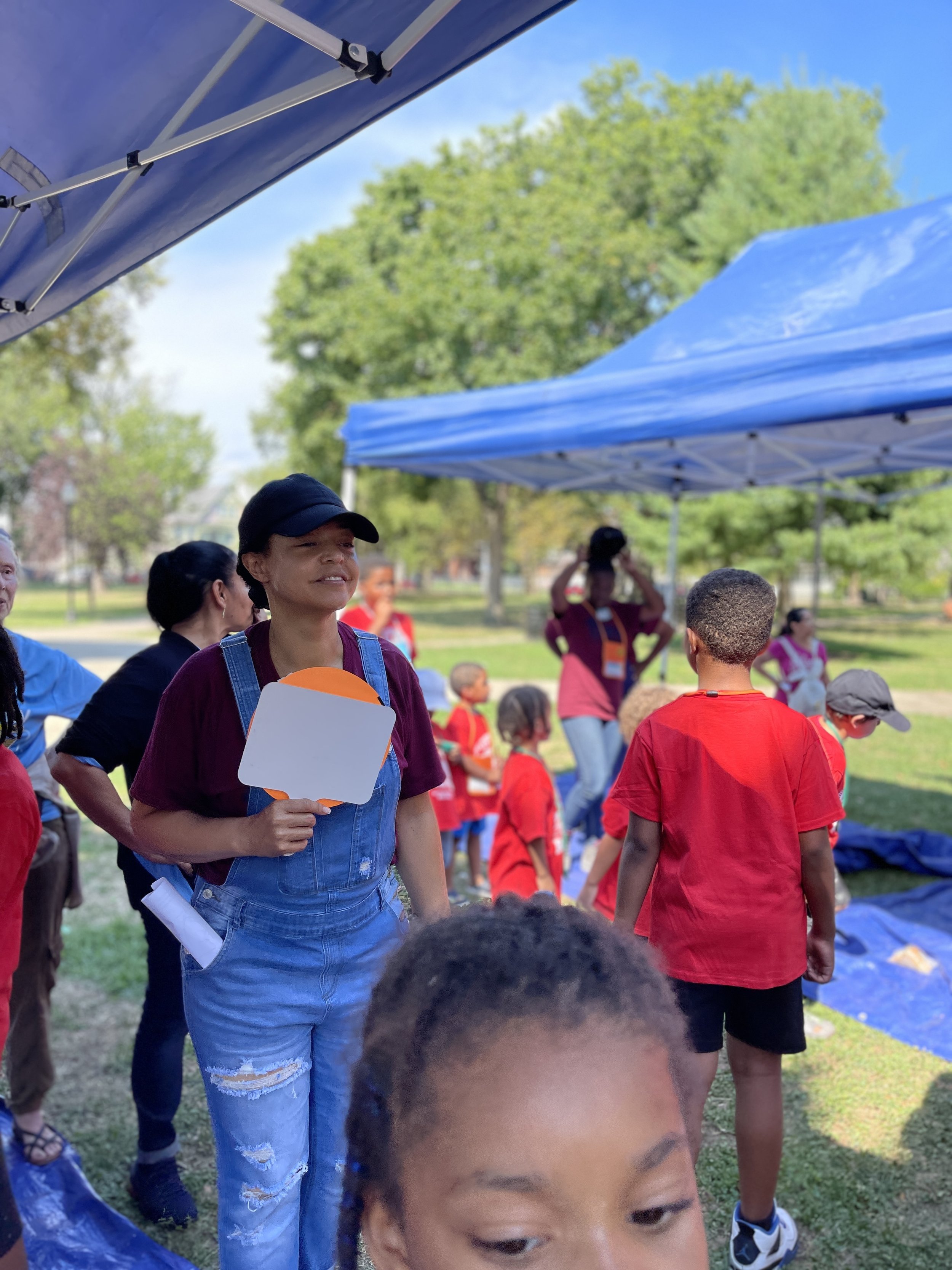 A group of people, including children and adults, gathering outdoors under blue canopies in a park on a sunny day. Some children are wearing red shirts and there are green trees in the background.