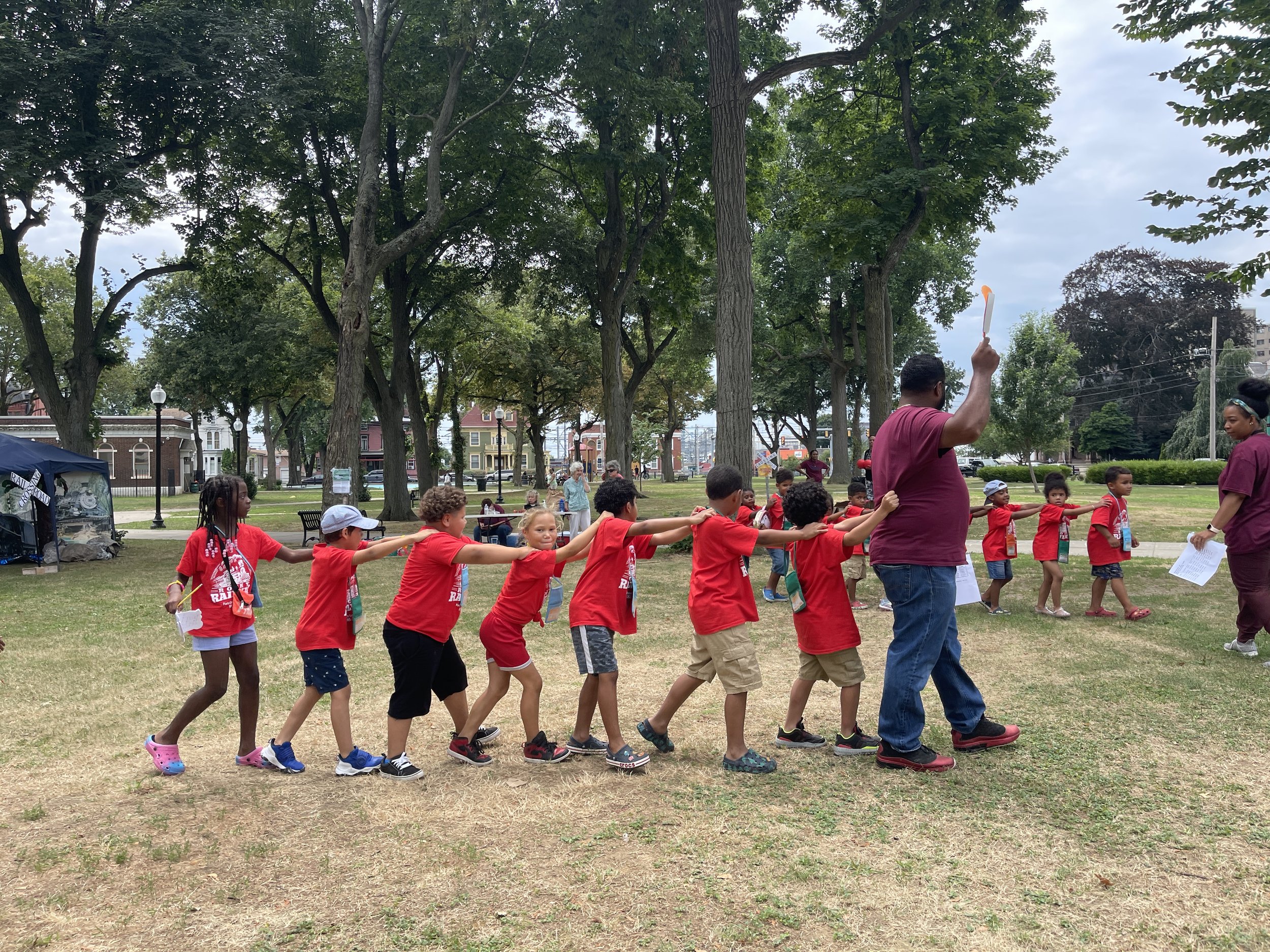 Children in red shirts and hats playing tug of war in a park, guided by two adults, under a large tree with a cityscape visible in the background.