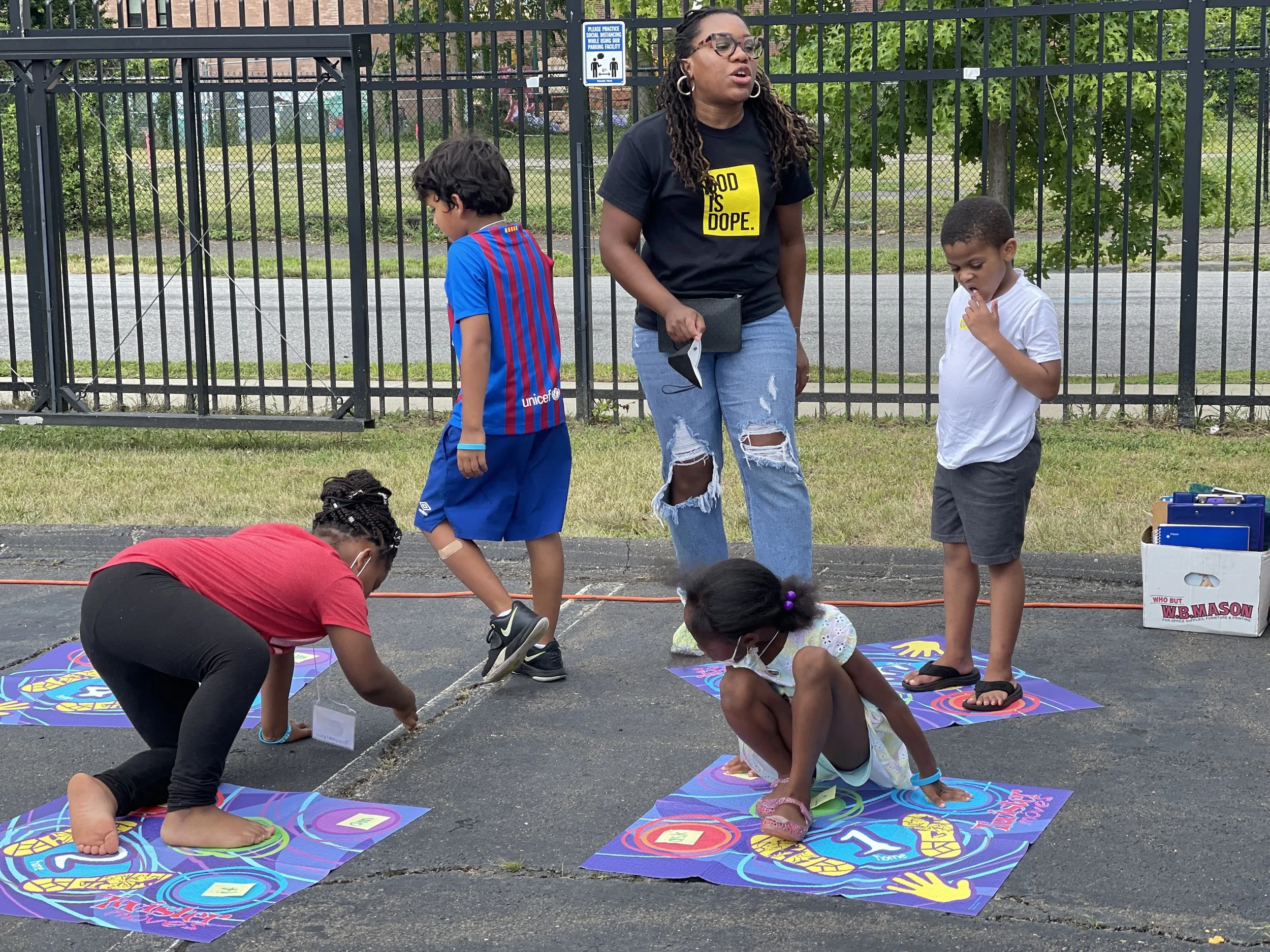 Children playing a large, colorful game on the pavement with an adult supervising nearby, outdoors next to a black fence and grassy area.