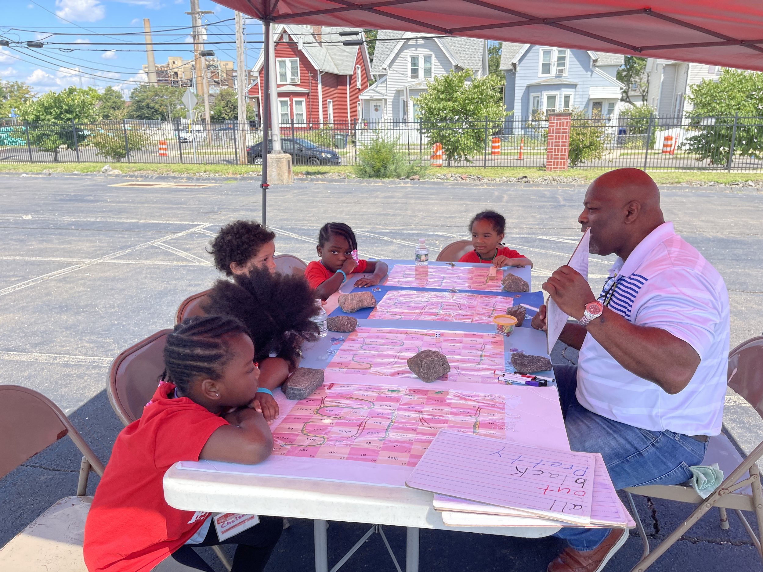 Boy teaching children spelling words with letter tiles at an outdoor table with rocks and colorful decorations under a red canopy.