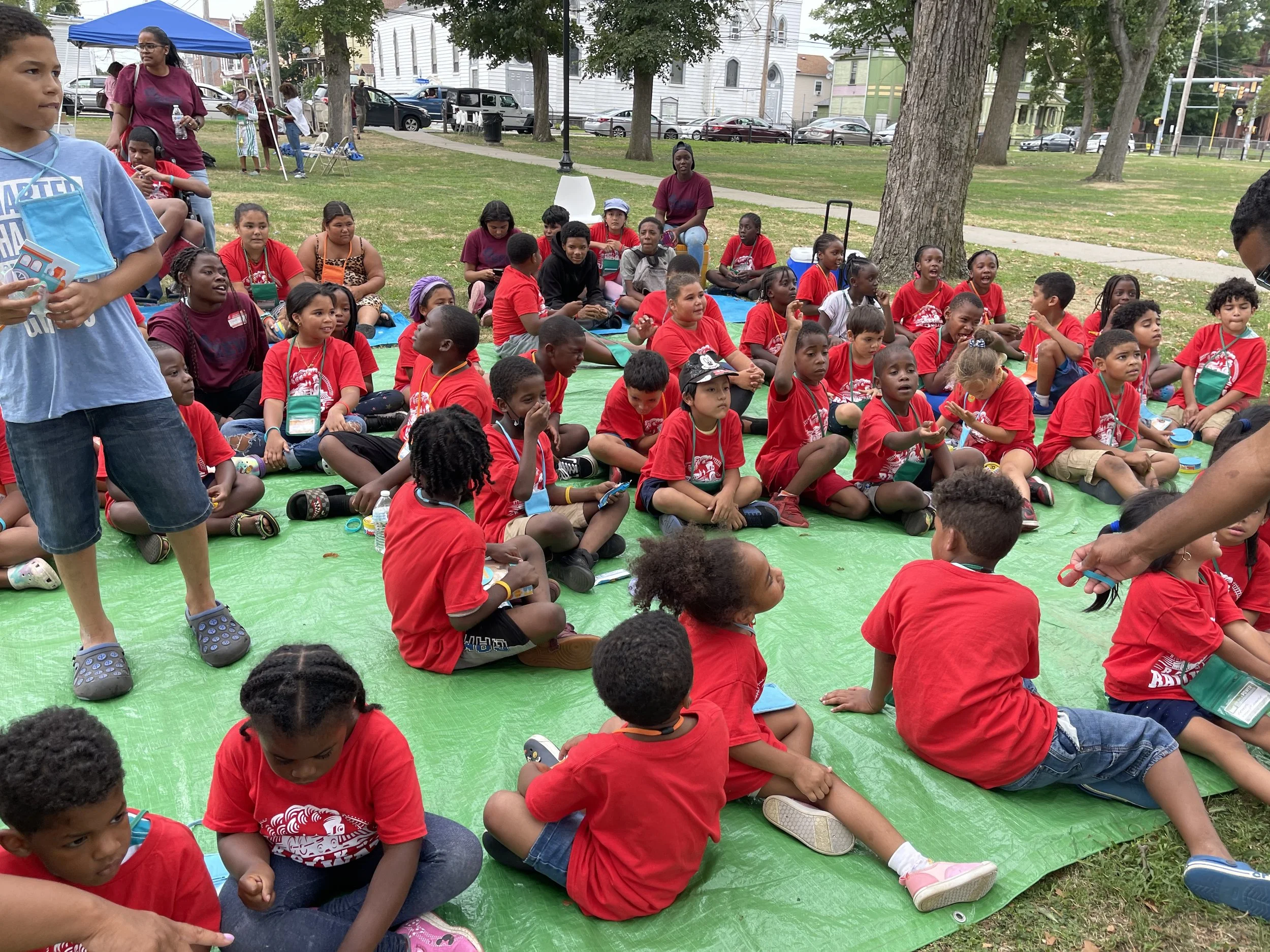 Children and adults sitting on a green tarp in a park, participating in an outdoor event on a sunny day, some wearing red shirts.