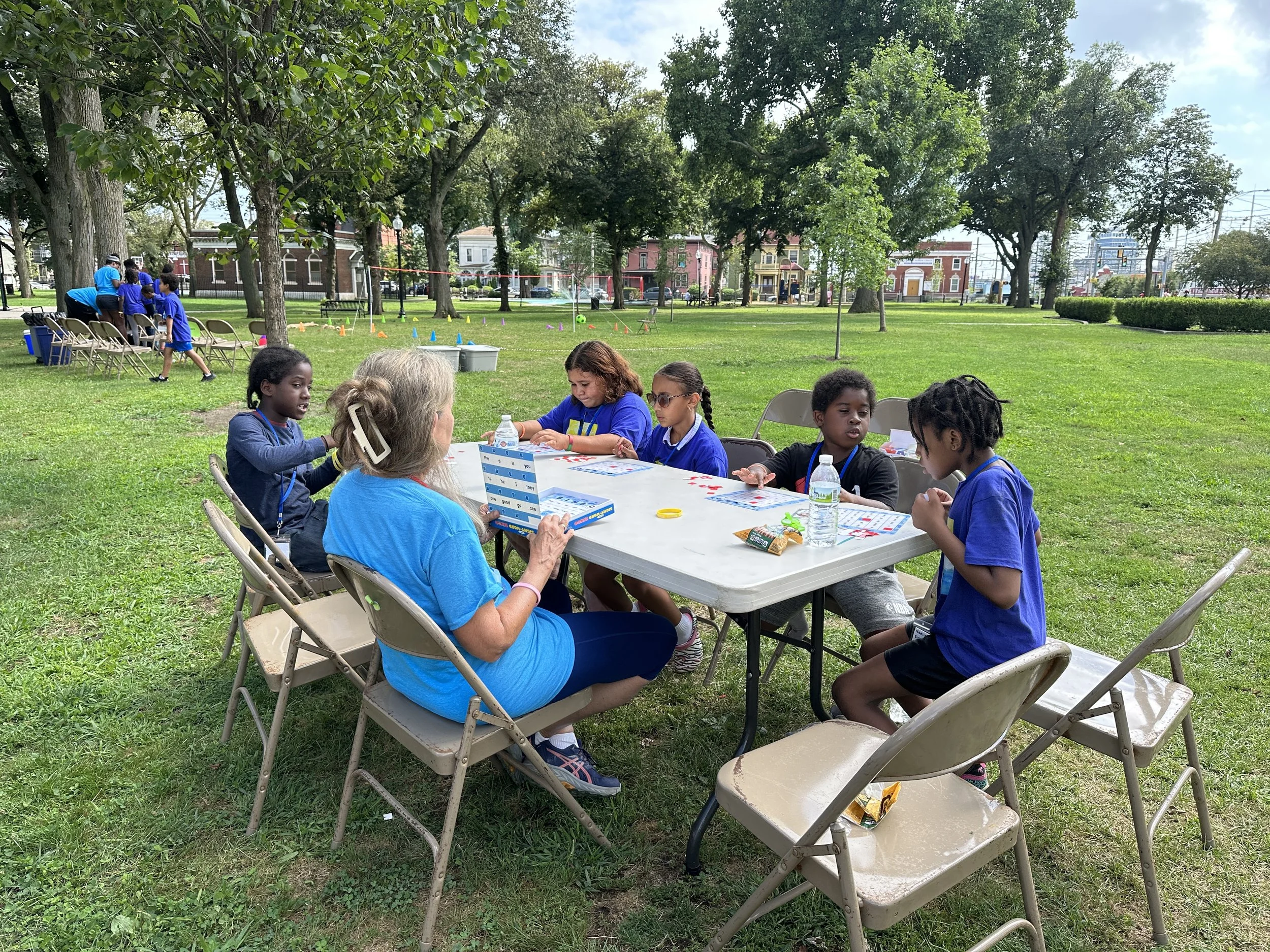 Group of children and an adult sitting at a white table outdoors in a park with green grass and trees, playing a game, with other children visible in the background near play equipment.