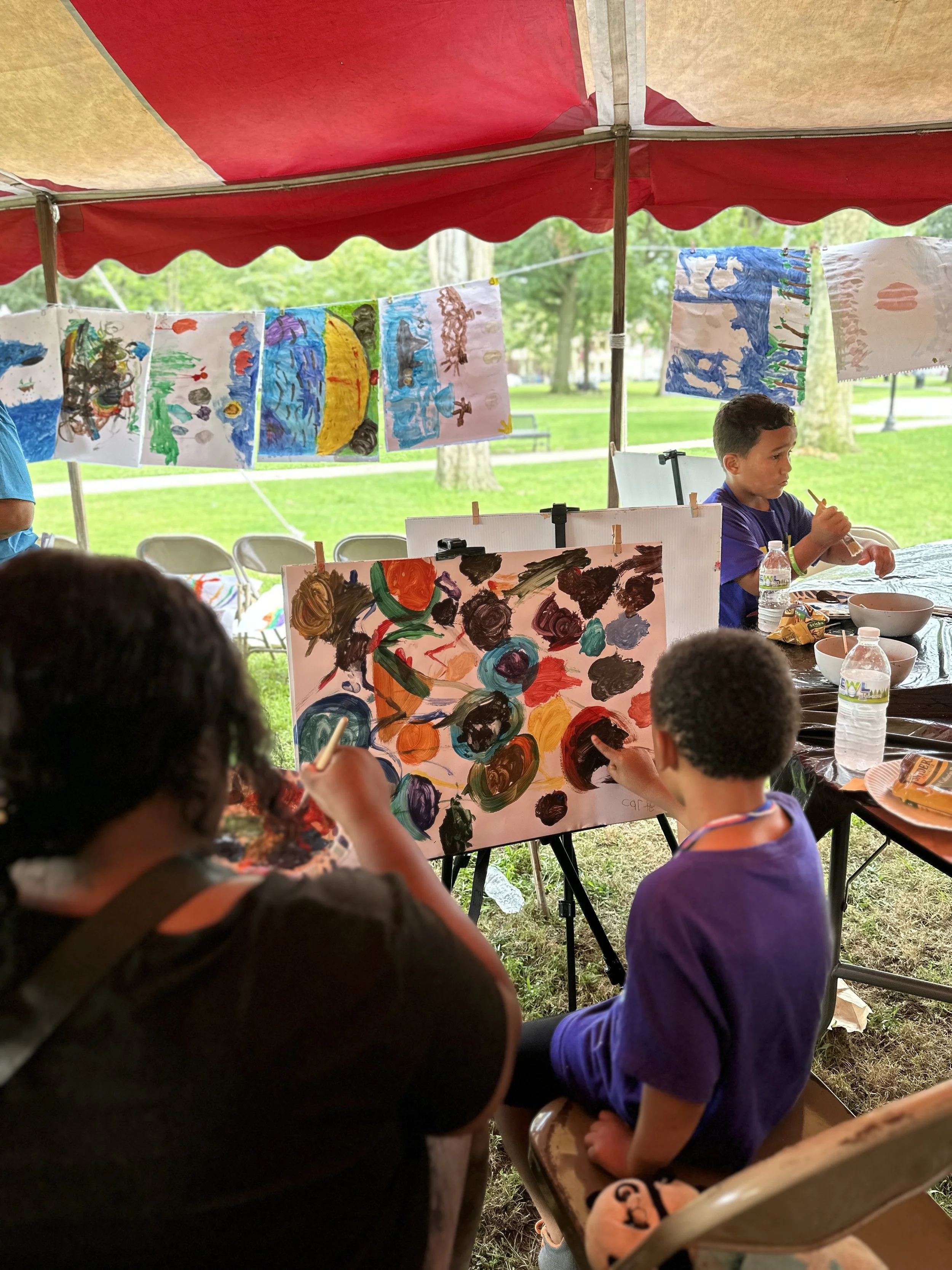 Children and adults painting colorful artwork on paper and display hanging under a red canopy in a park.