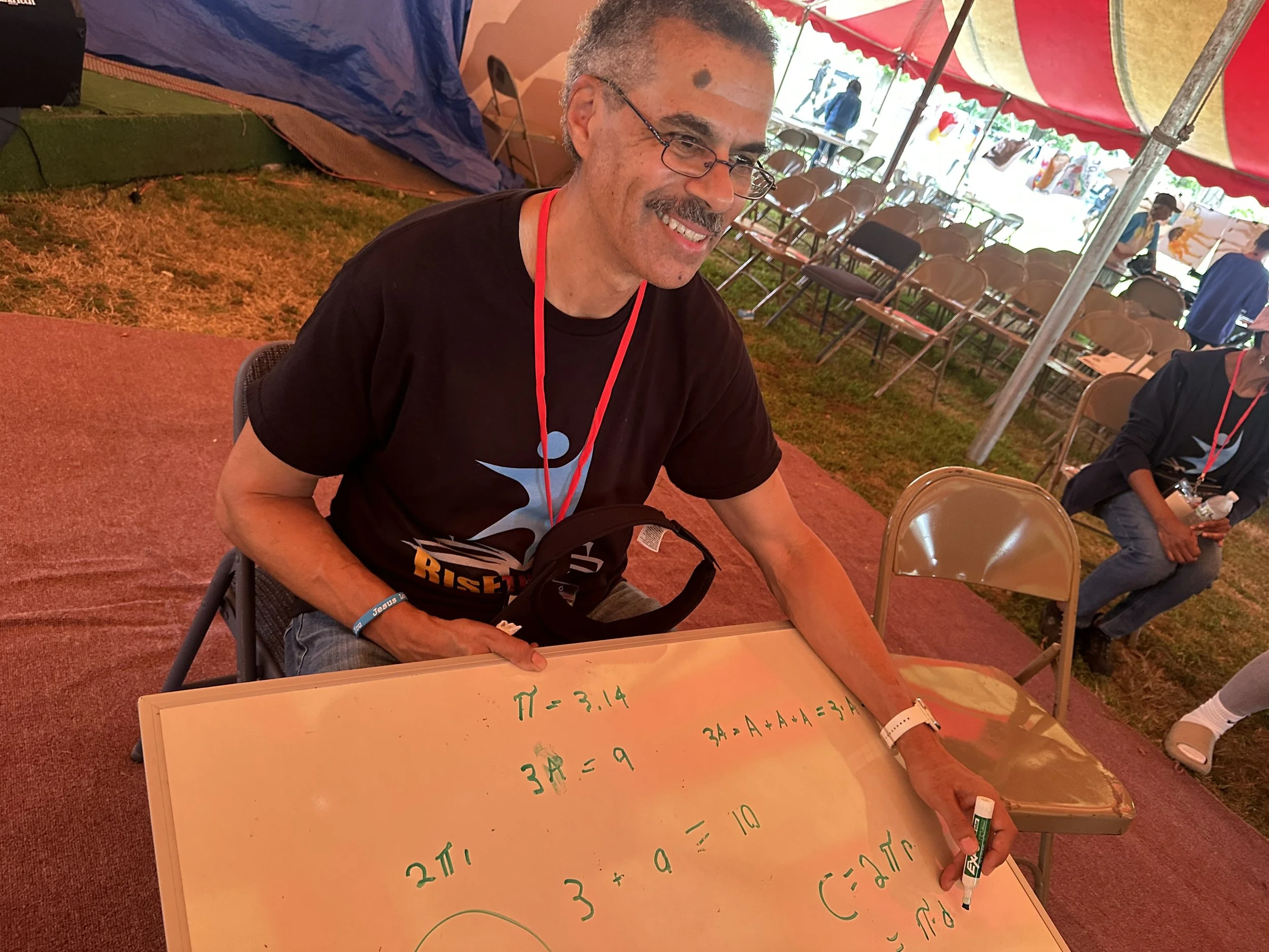 A smiling man with glasses, gray hair, and a mustache is sitting at a table inside a tent, writing mathematical formulas on a whiteboard with a green marker. The formulas include Pi, the area of a circle, and a volume calculation. There are empty cha