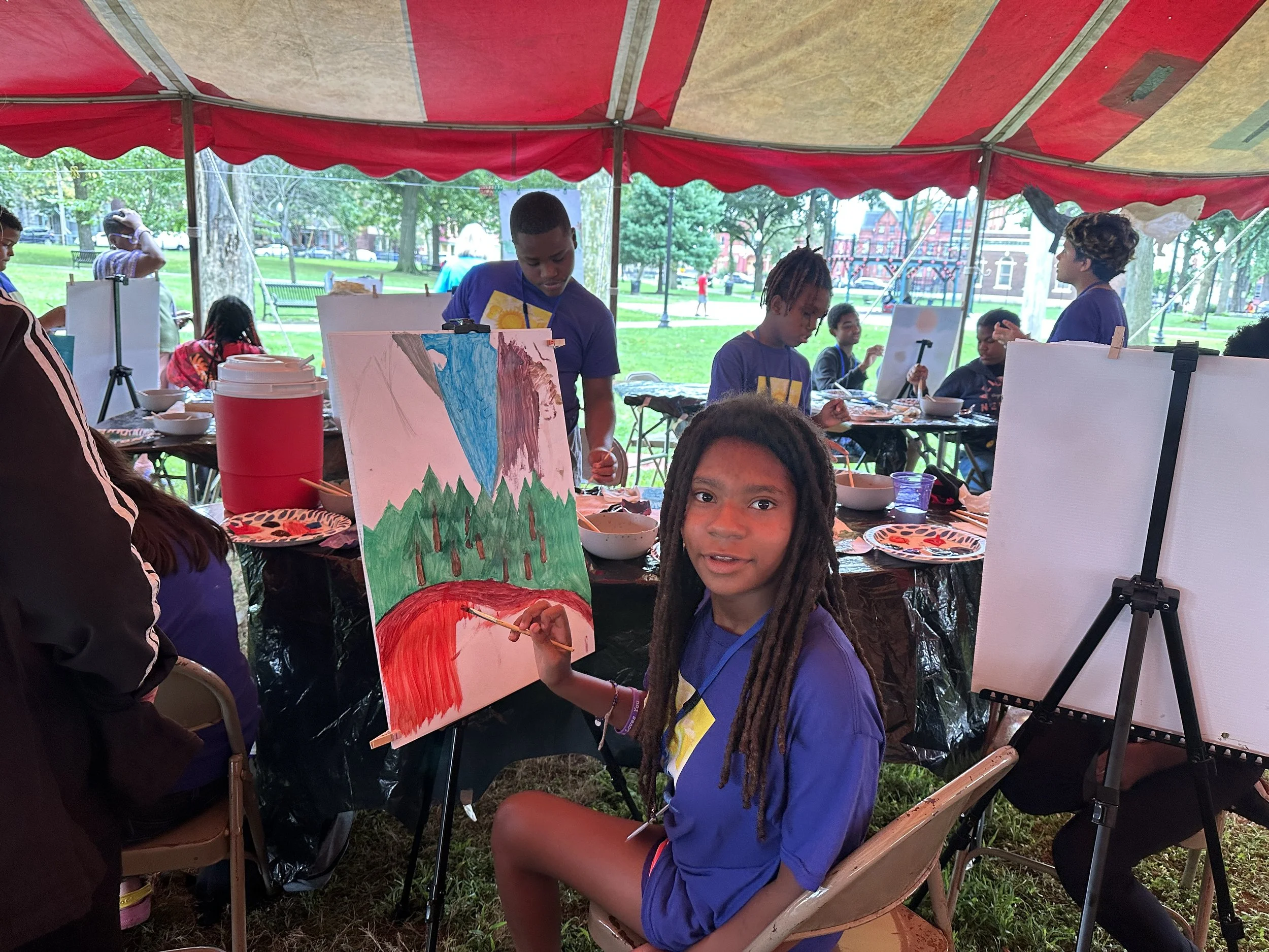 A young girl with dreadlocks sitting in front of a painting of a mountain and trees under a blue sky, holding a paintbrush, in an outdoor art activity under a red and beige tent.
