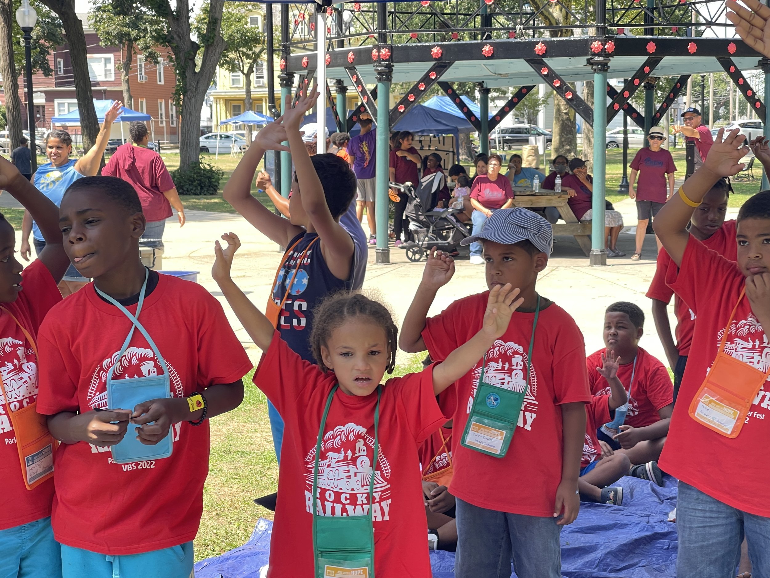 Children wearing red shirts participating in an outdoor event, raising their hands, with adults and a slide in the background under a sunny sky.