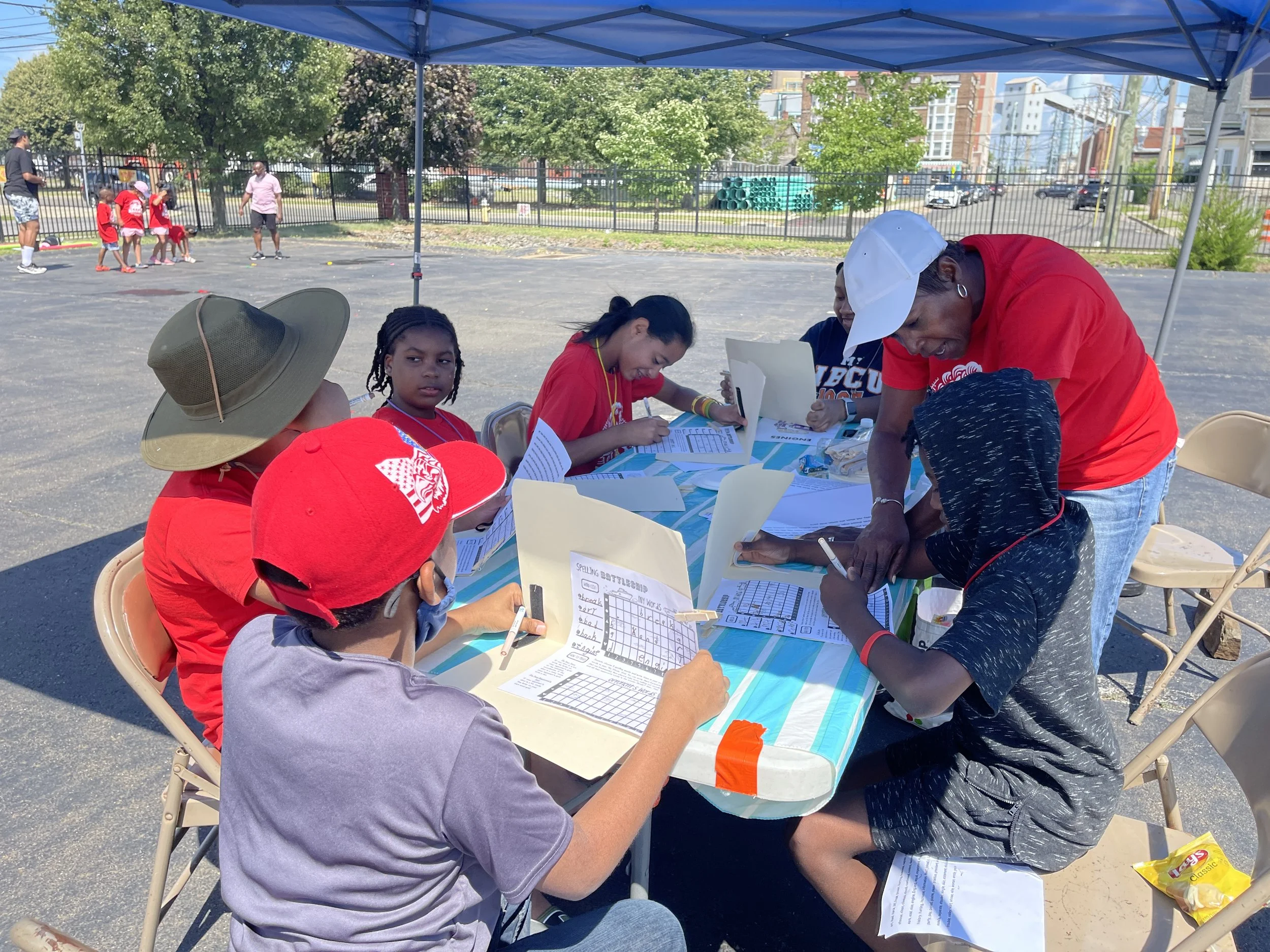 Children and adults seated at a table outside under a canopy, working on papers and activities, while others play in the background on a sunny day.