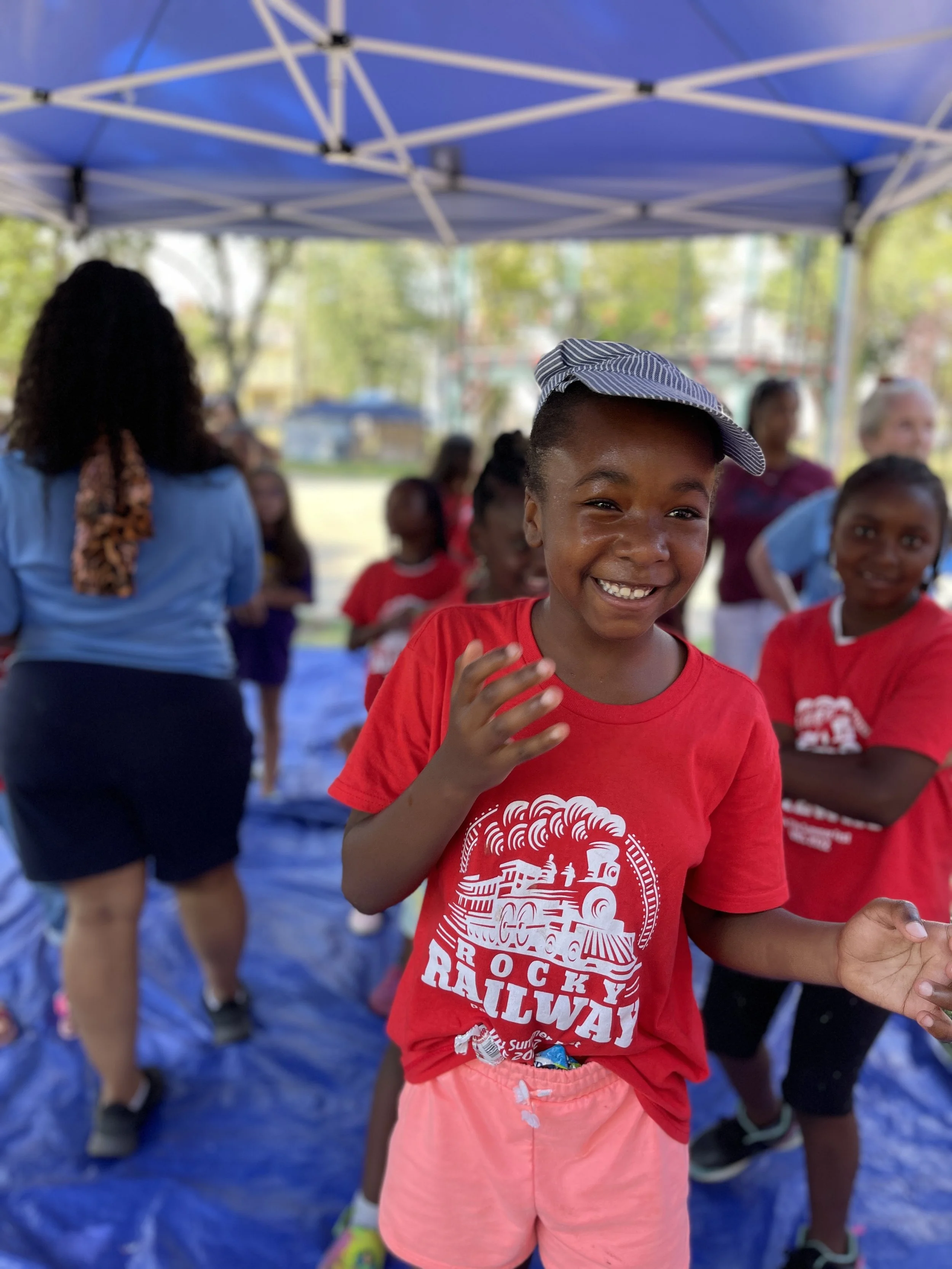 A young girl smiling and playing under a blue canopy tent with other children and adults in the background, wearing a red t-shirt with a railway train graphic and pink shorts.