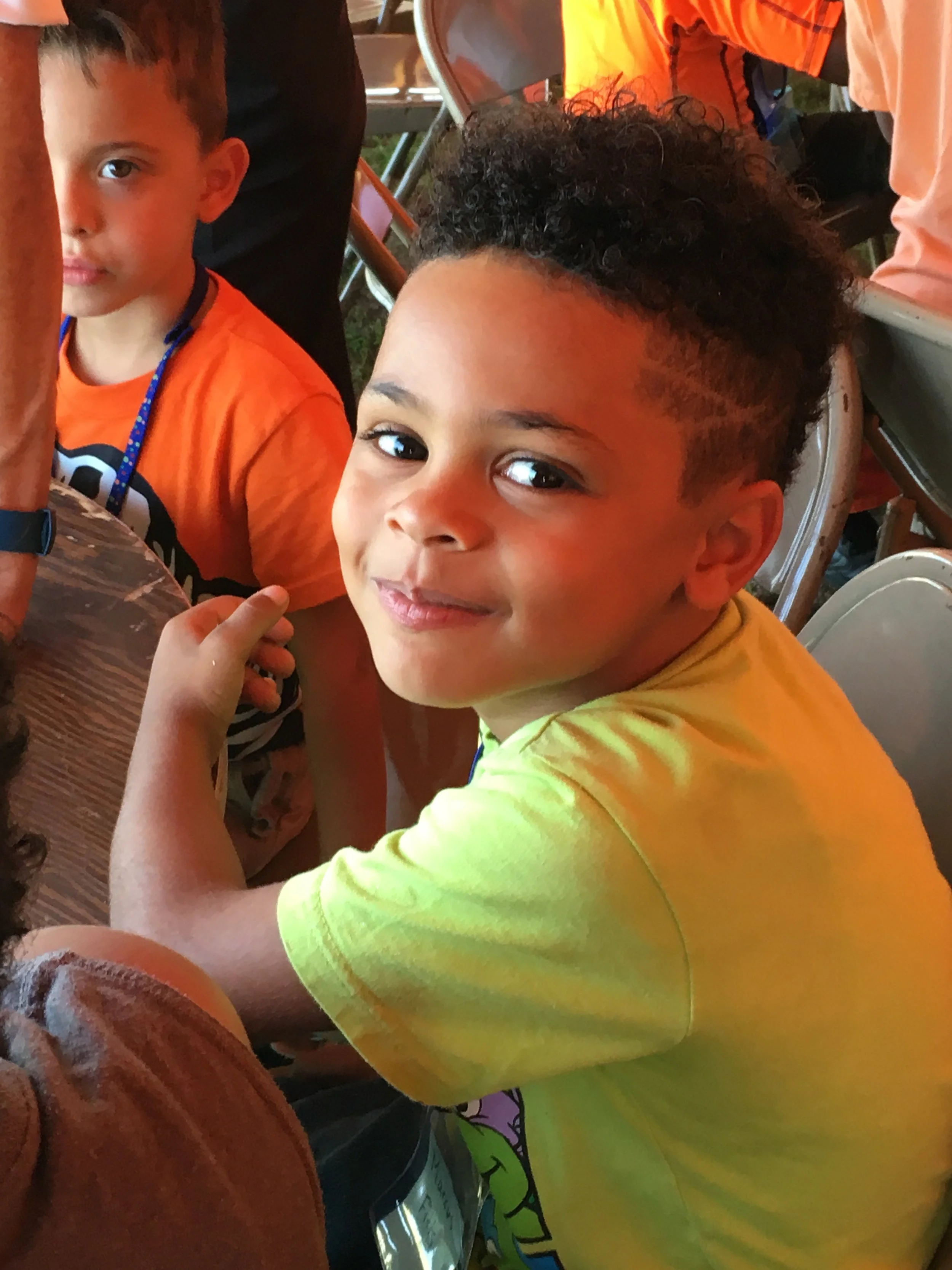 Young boy with curly dark hair, wearing a yellow t-shirt, sitting at a table, looking up at the camera with a slight smile.