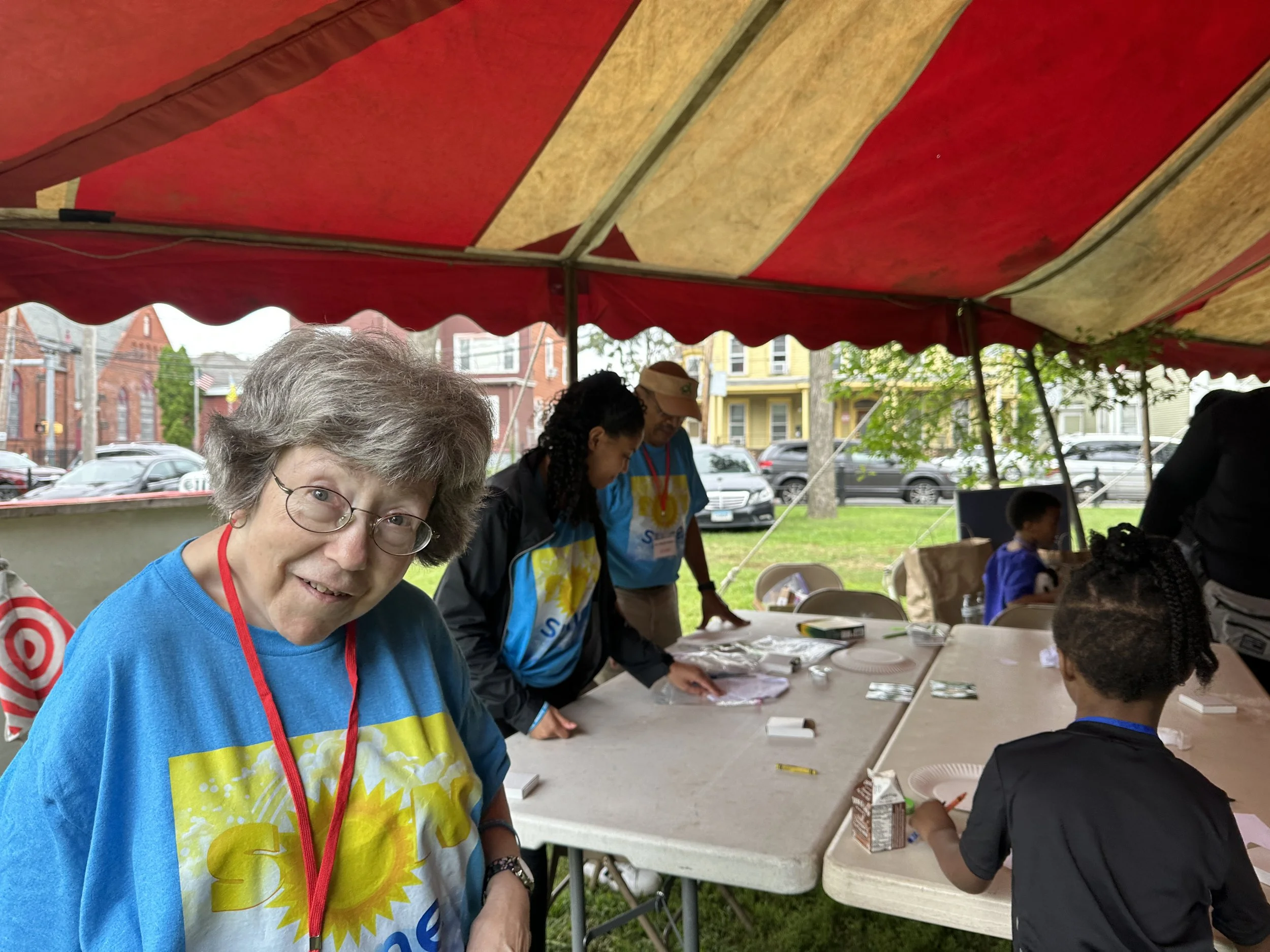 A group of people under a red and yellow striped tent, engaged in activities at a long table outdoors, with a woman wearing glasses and a blue T-shirt smiling at the camera.