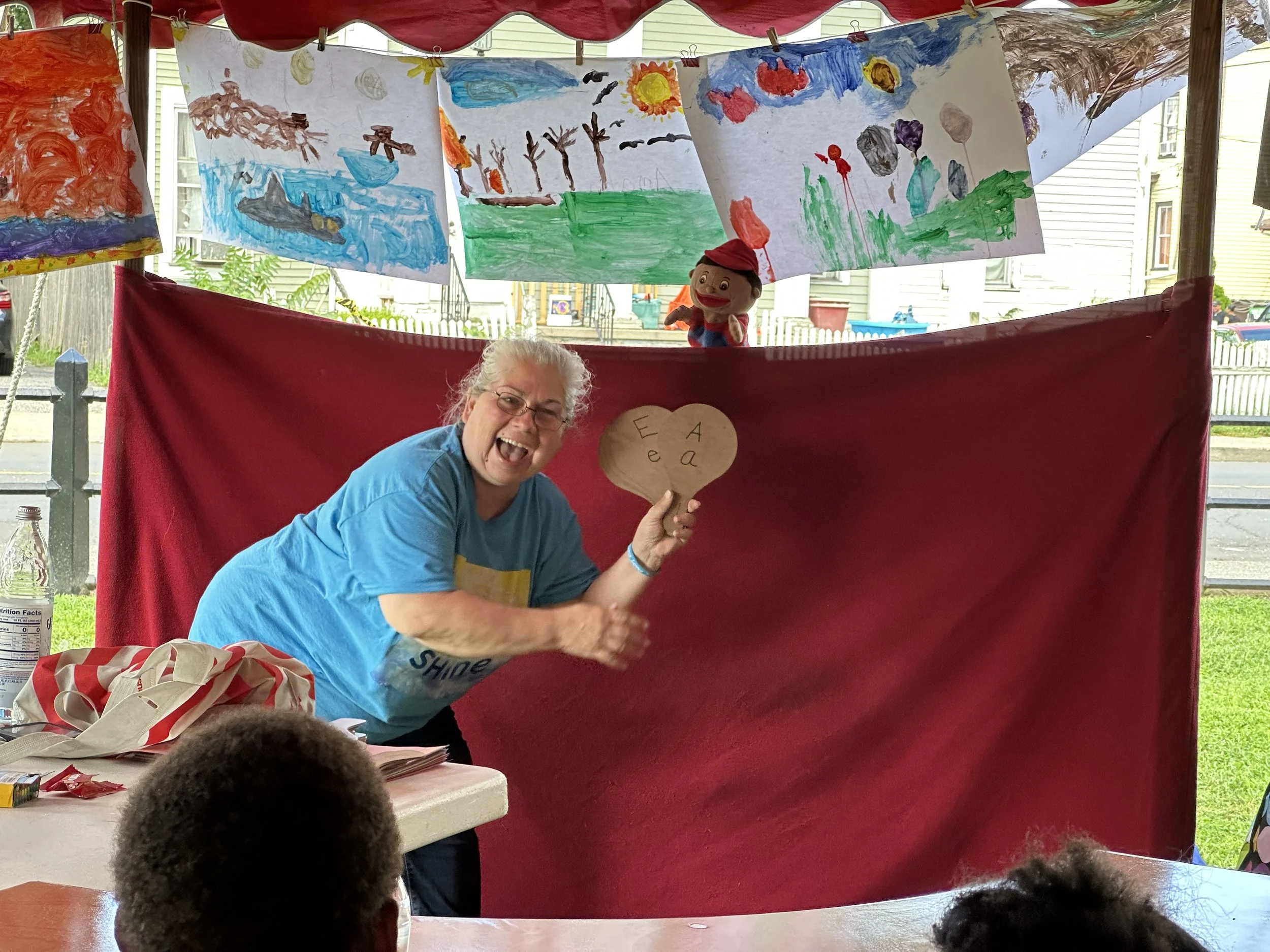 A woman with gray hair and glasses is smiling and holding a cardboard heart with the letters E, A, and A written on it. She is standing in front of a red fabric backdrop with children's artwork hanging above. The setting appears to be a classroom or 