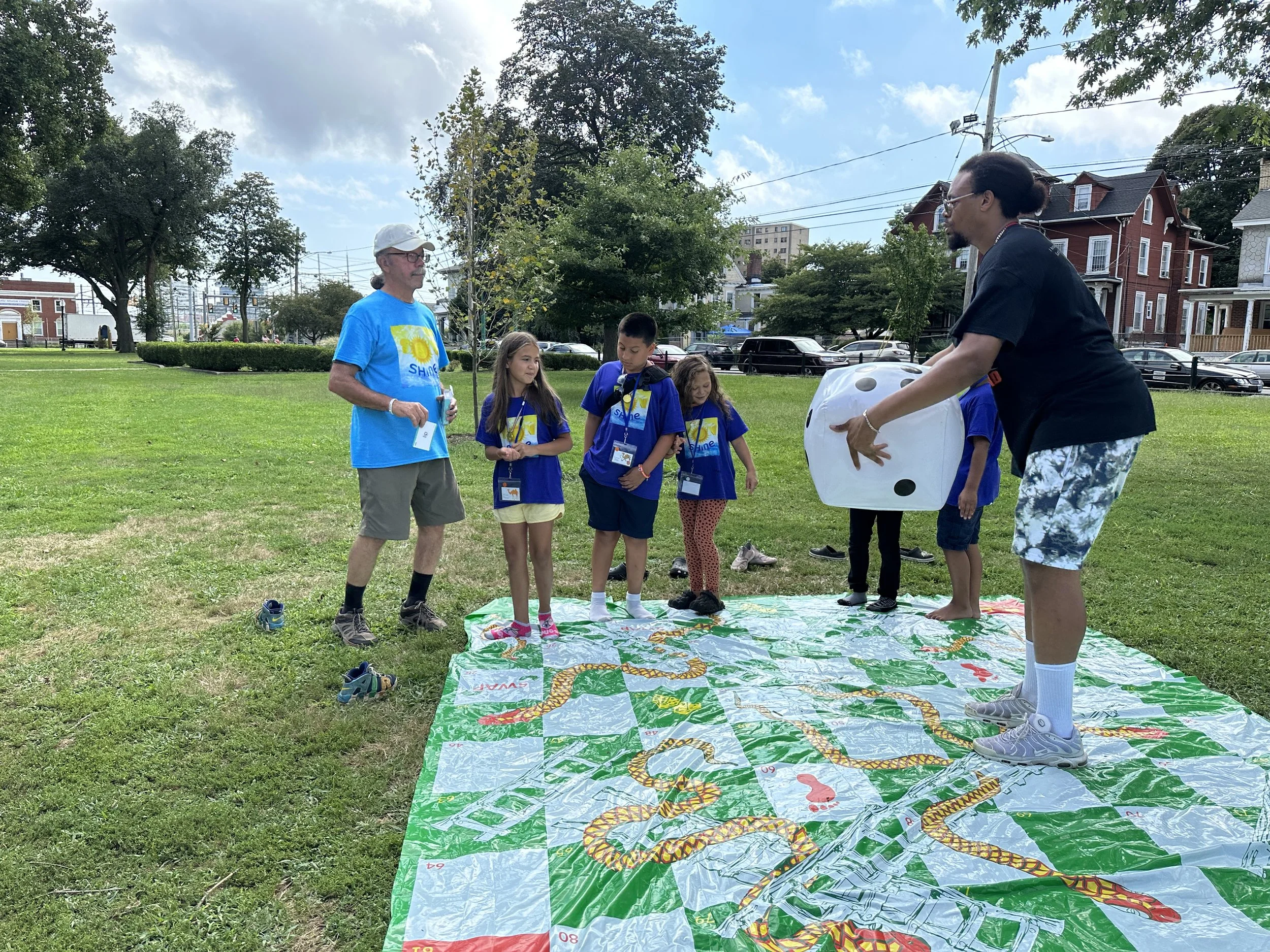Adults and children playing Twister on a grassy field during a sunny day, with houses and trees in the background.