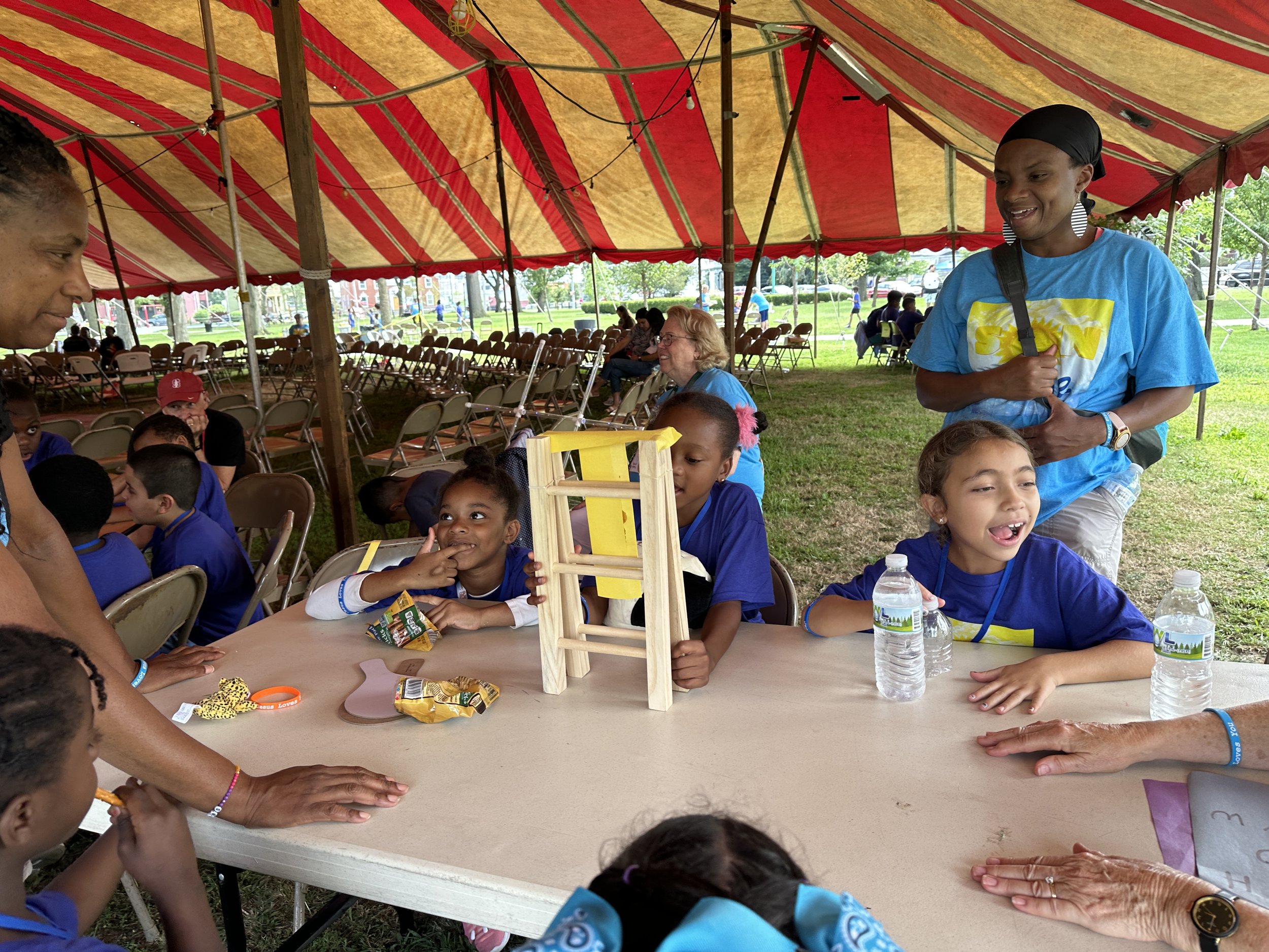 Children and adults sitting under a large red and yellow striped canopy, engaging in activities at a table outdoors on a grassy area, with more chairs and people visible in the background.