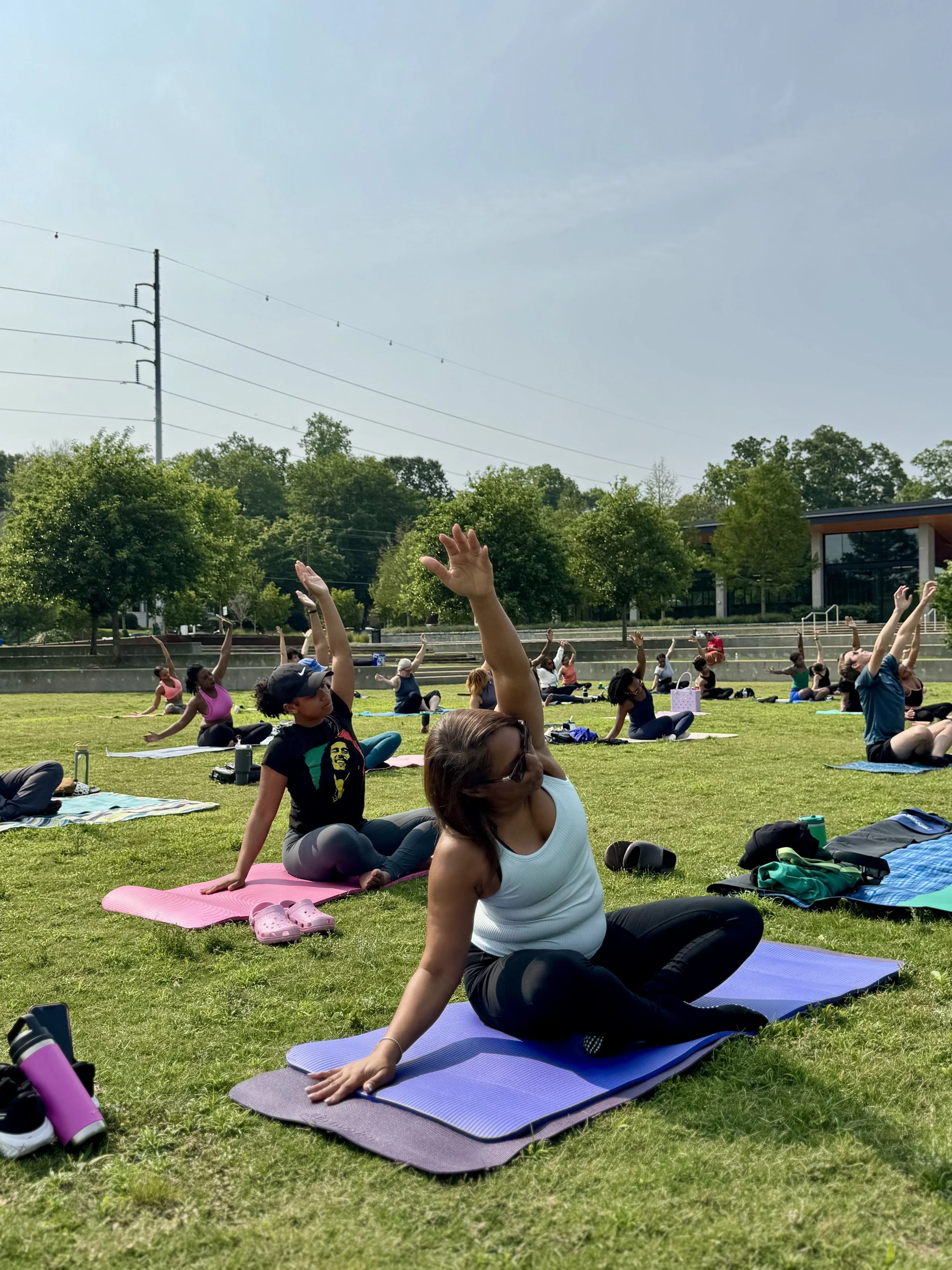People participating in a yoga class outdoors on a grassy field, practicing stretching poses on colorful mats during the day.