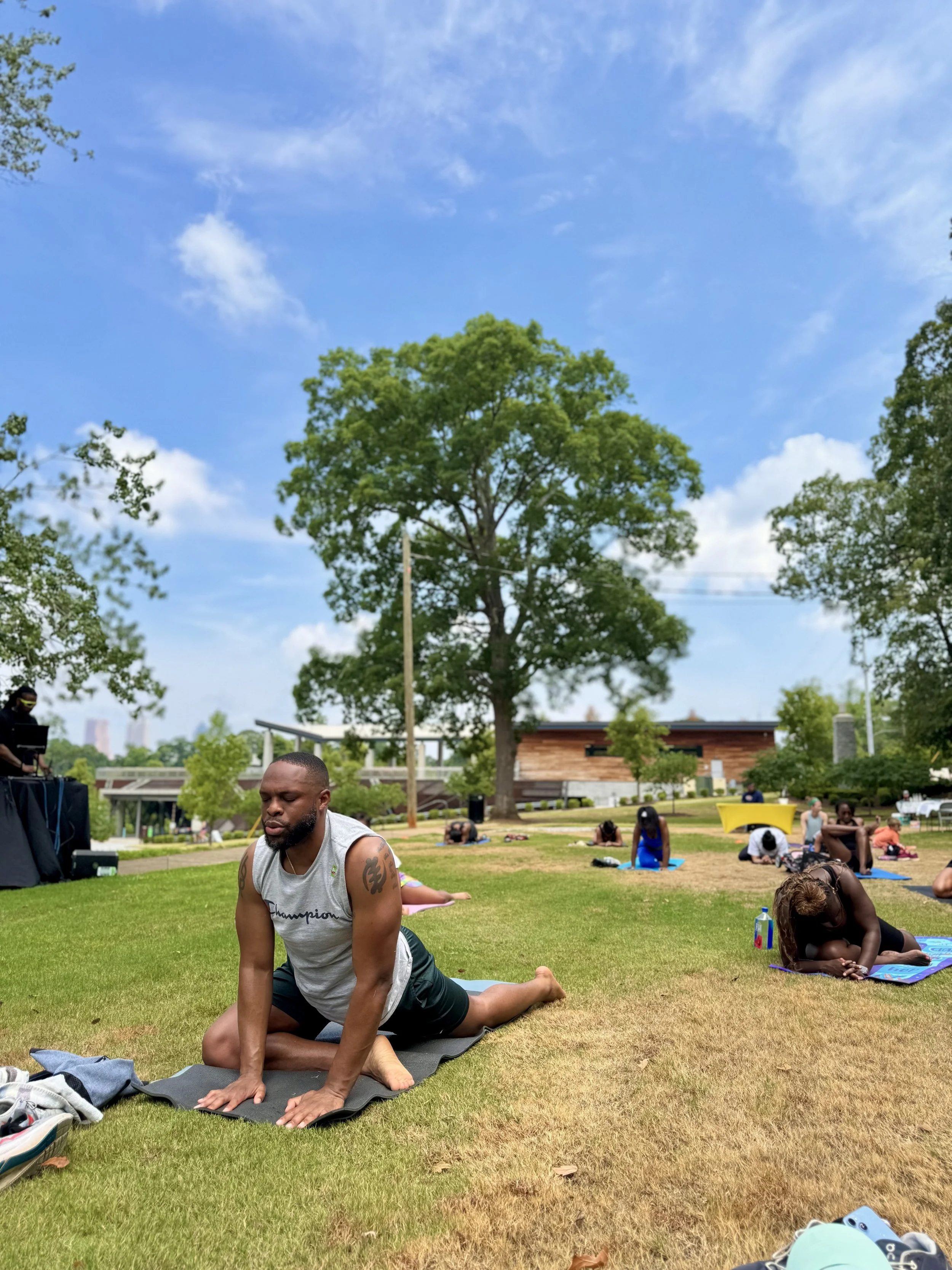 People practicing yoga outdoors on mats in a park on a sunny day with blue sky and large trees.