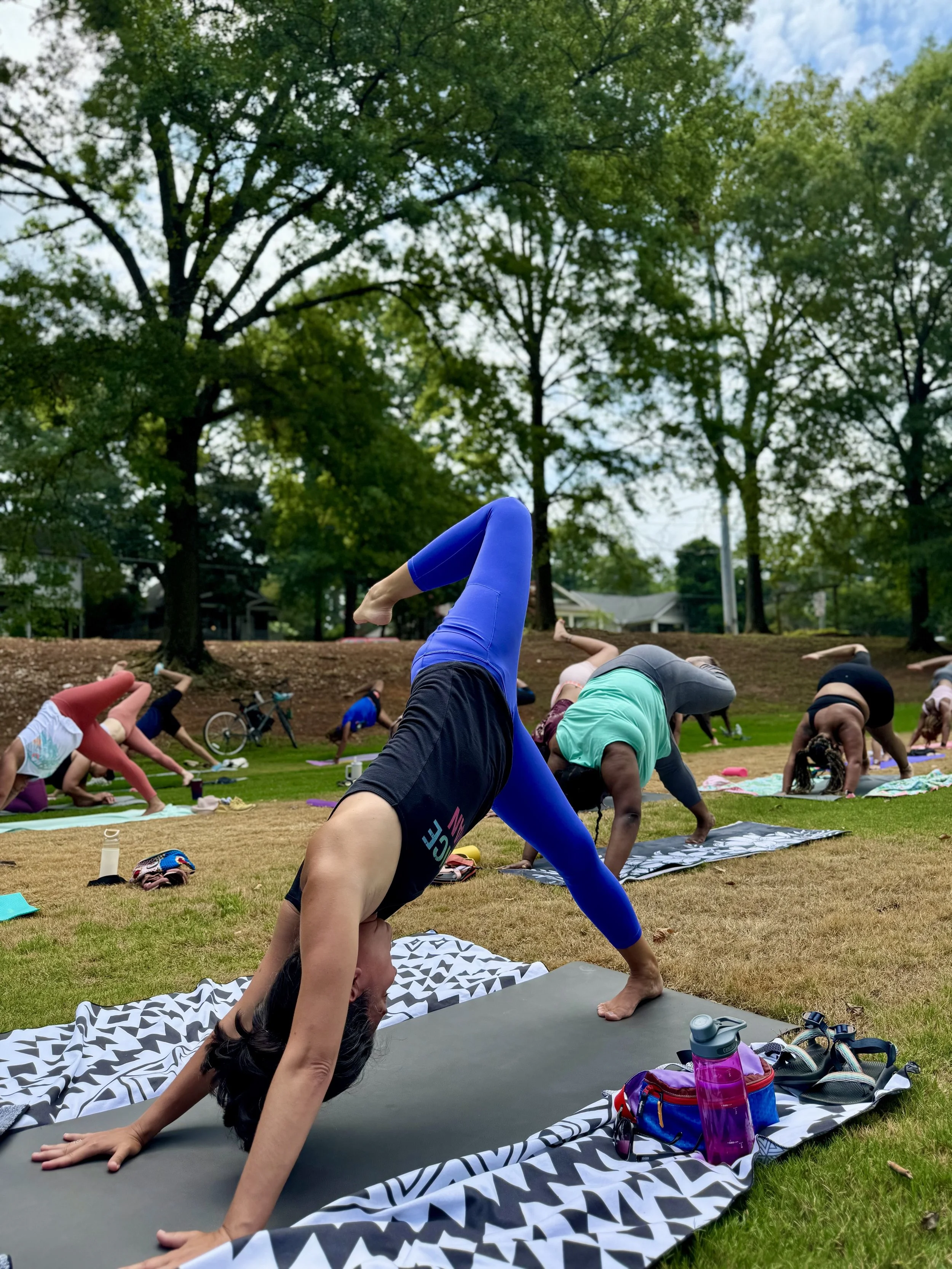 Group of people practicing yoga outdoors on mats in a park during daytime, with trees and blue sky in the background.