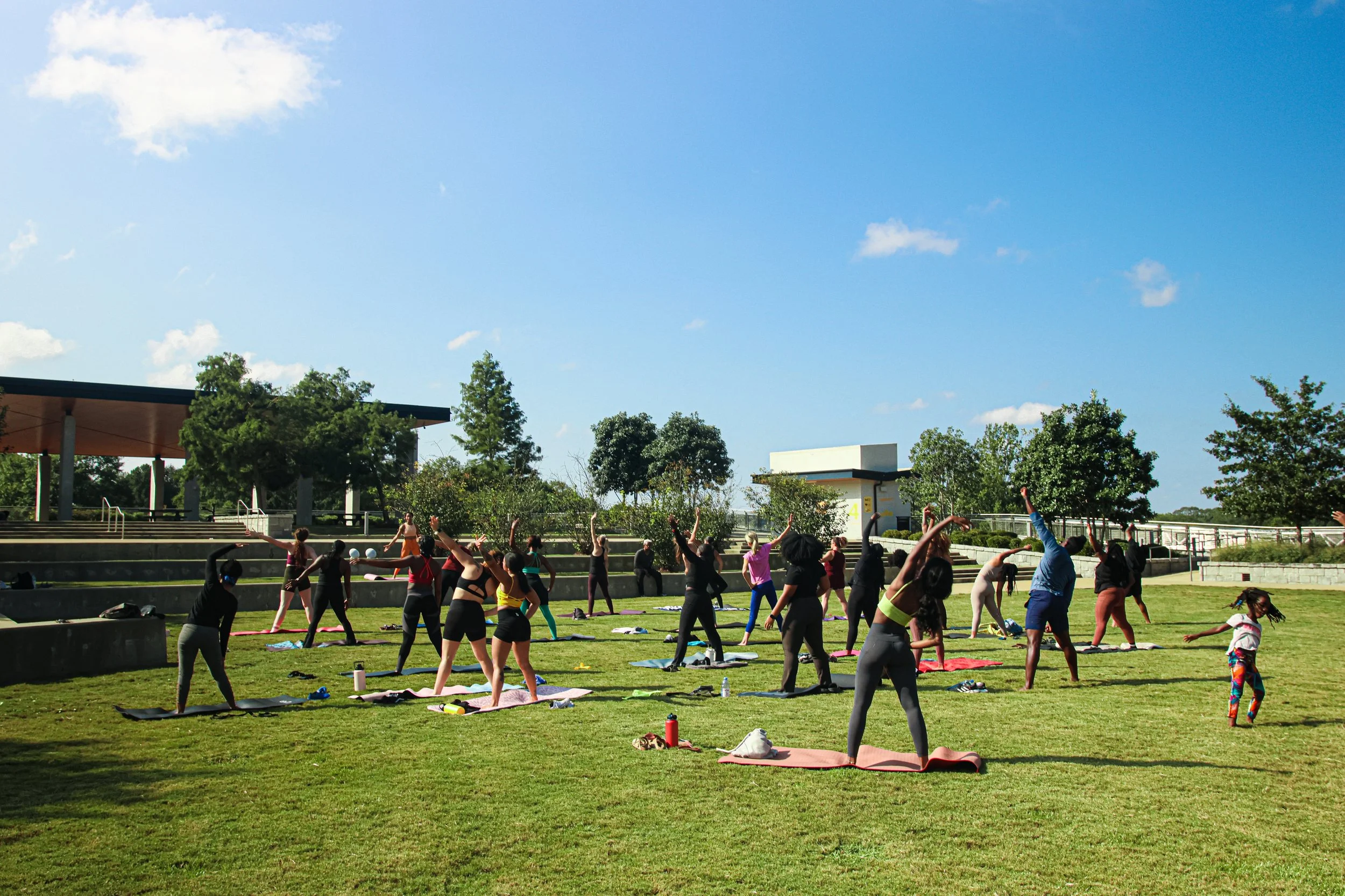 Group of people participating in an outdoor Pilates in the Park class on a grassy field on a sunny day with a blue sky and trees in the background.