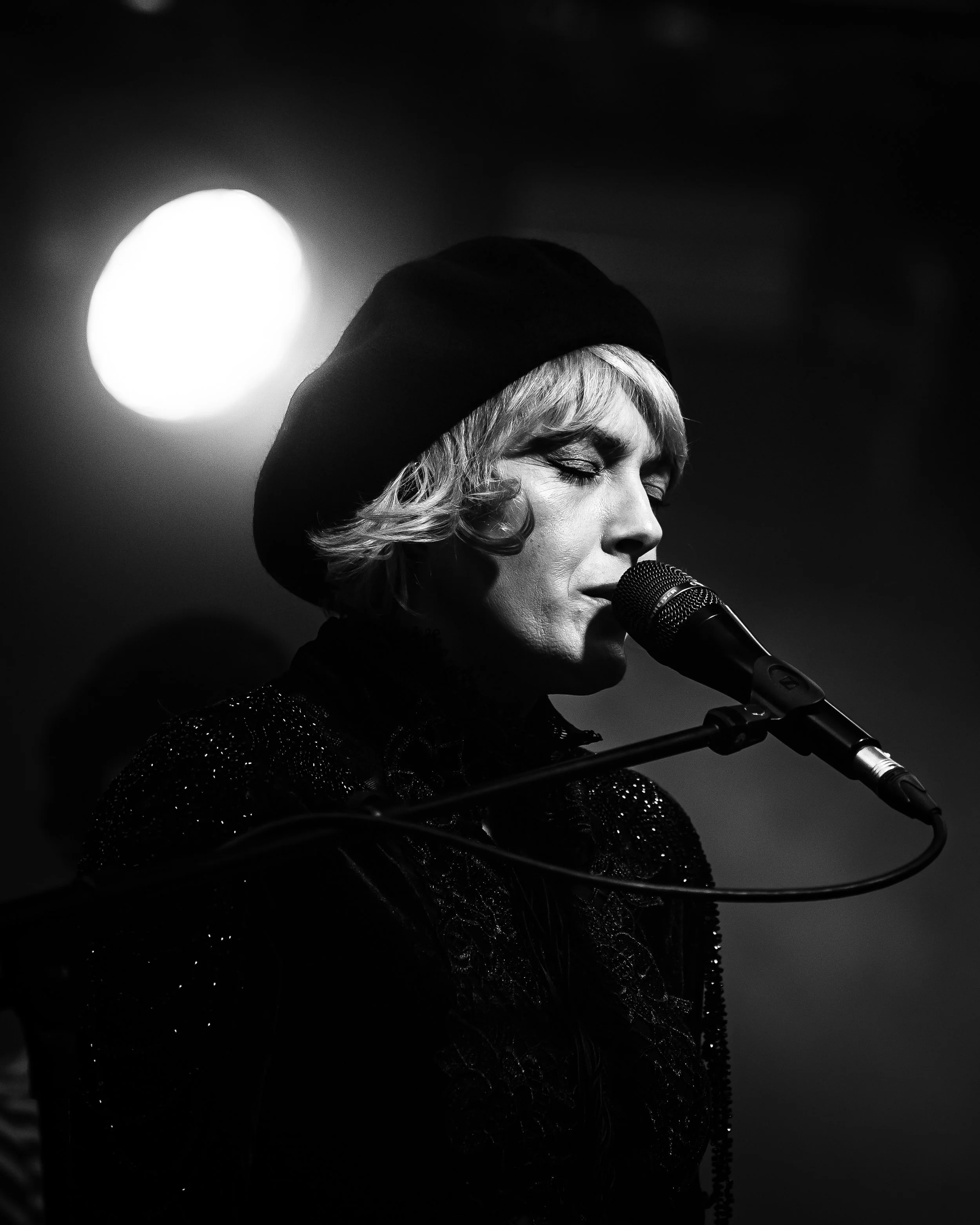 A woman singing into a microphone, wearing a hat and a sparkly outfit, with a large bright light in the background.