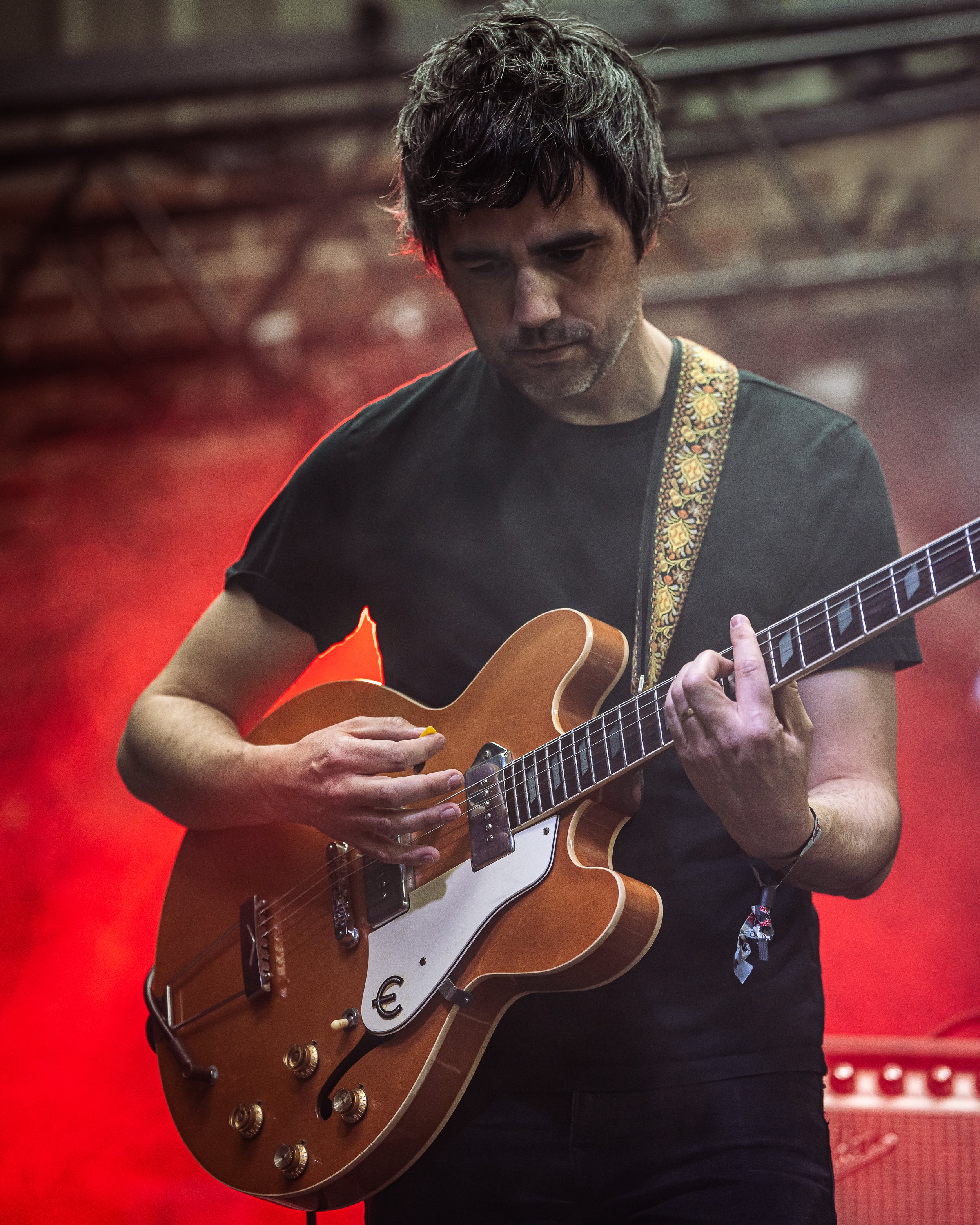 A man with dark, slightly messy hair is playing an electric guitar with a sunburst finish in front of a red background. He appears focused on his playing, and is wearing a black t-shirt and a patterned guitar strap.